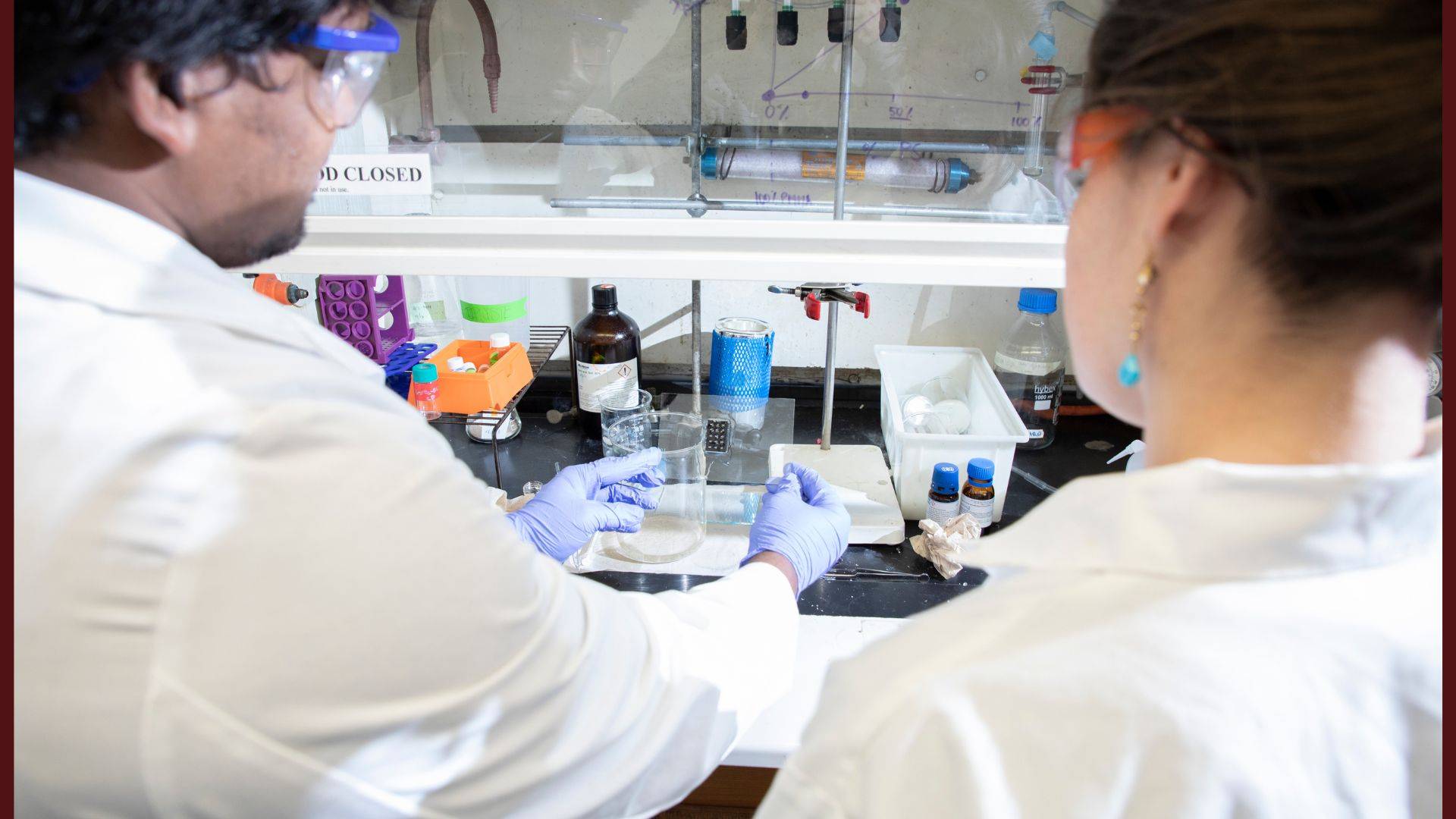Two students wearing lab coats, gloves, and safety goggles work together at a fume hood in a chemistry lab. One holds a beaker while the other observes, with various lab supplies, bottles, and equipment organized on the bench.