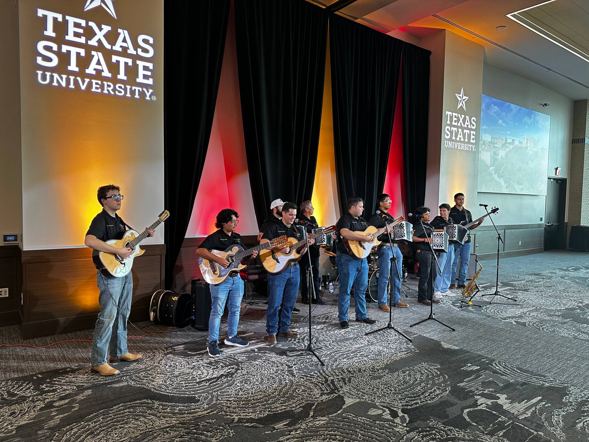 Band members wearing black polos and blue jeans hold guitars and accordions.