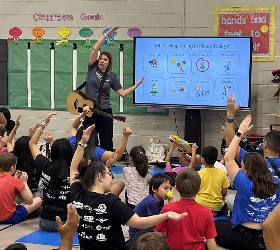 kids sit on floor facing a screen with image standing beside it holding an arm out