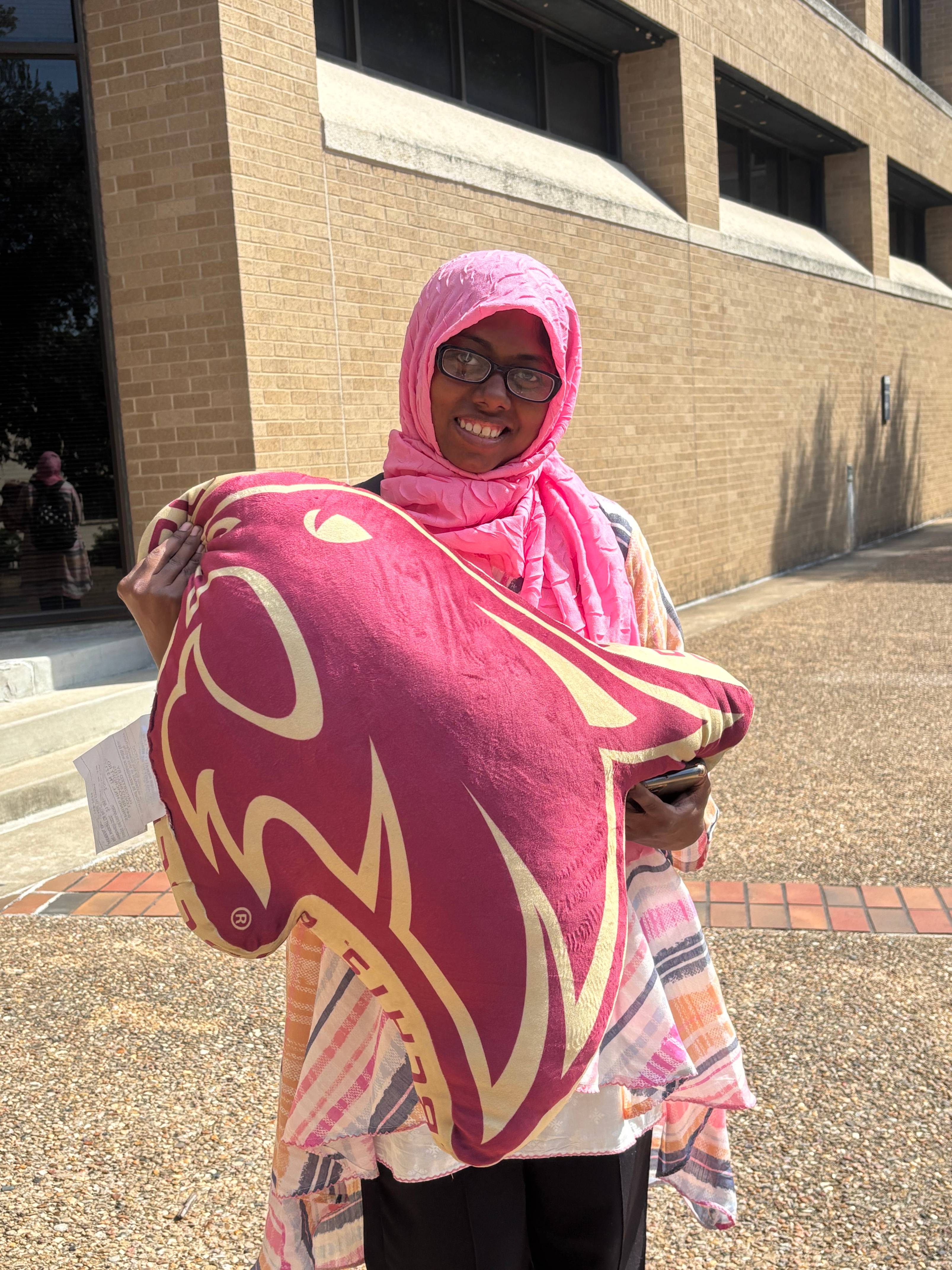 A smiling student wearing a pink hijab and glasses holds a large, maroon and gold pillow shaped like the Texas State Bobcat logo. They stand outside a tan brick building on a sunny day.