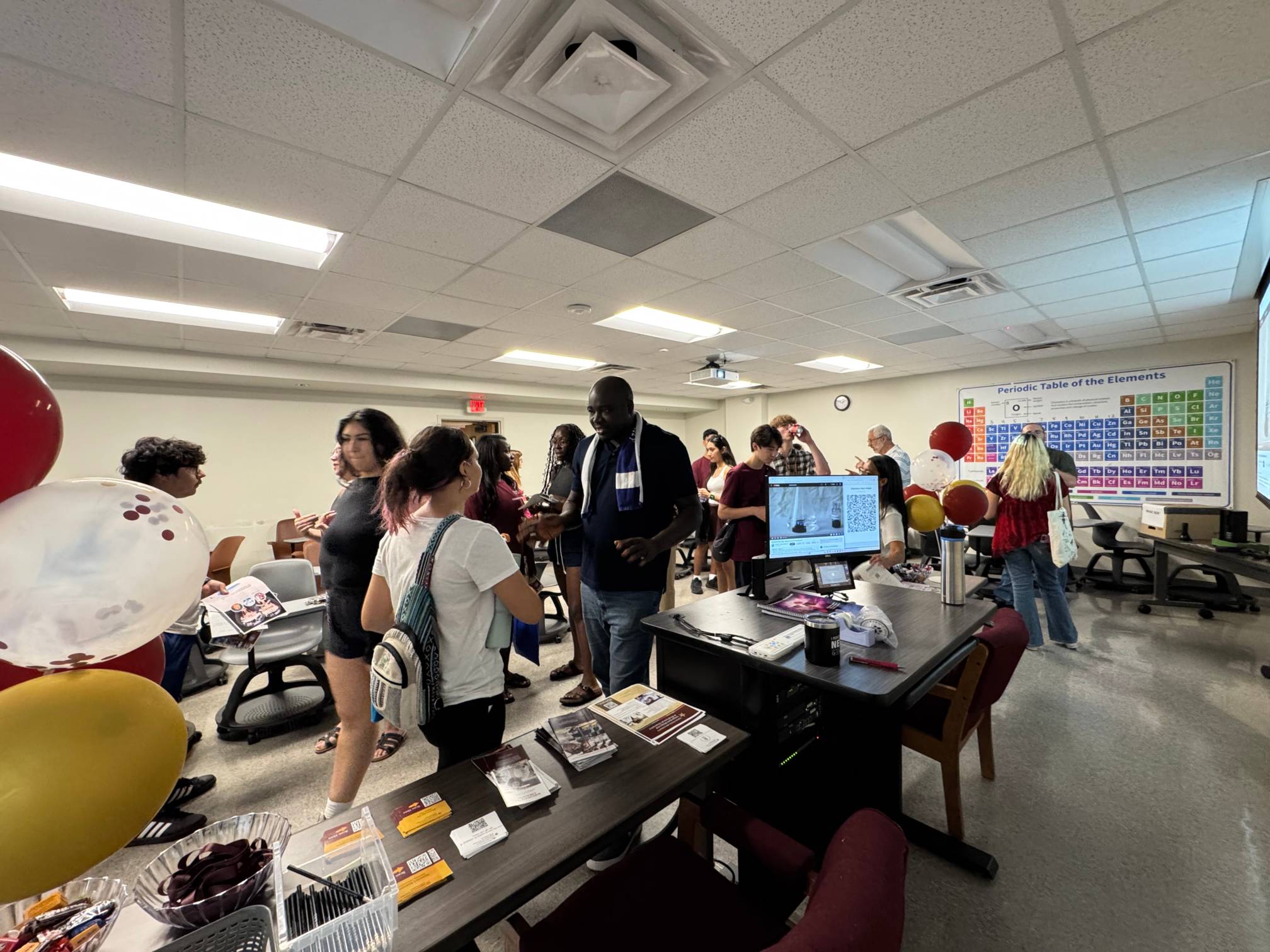 A wide-angle shot of a group of people gathered in a classroom. On the right, a large Periodic Table of the Elements chart is visible on the wall. Balloons, a table with refreshments, and a computer monitor are in the foreground, and people are standing and talking in small groups.