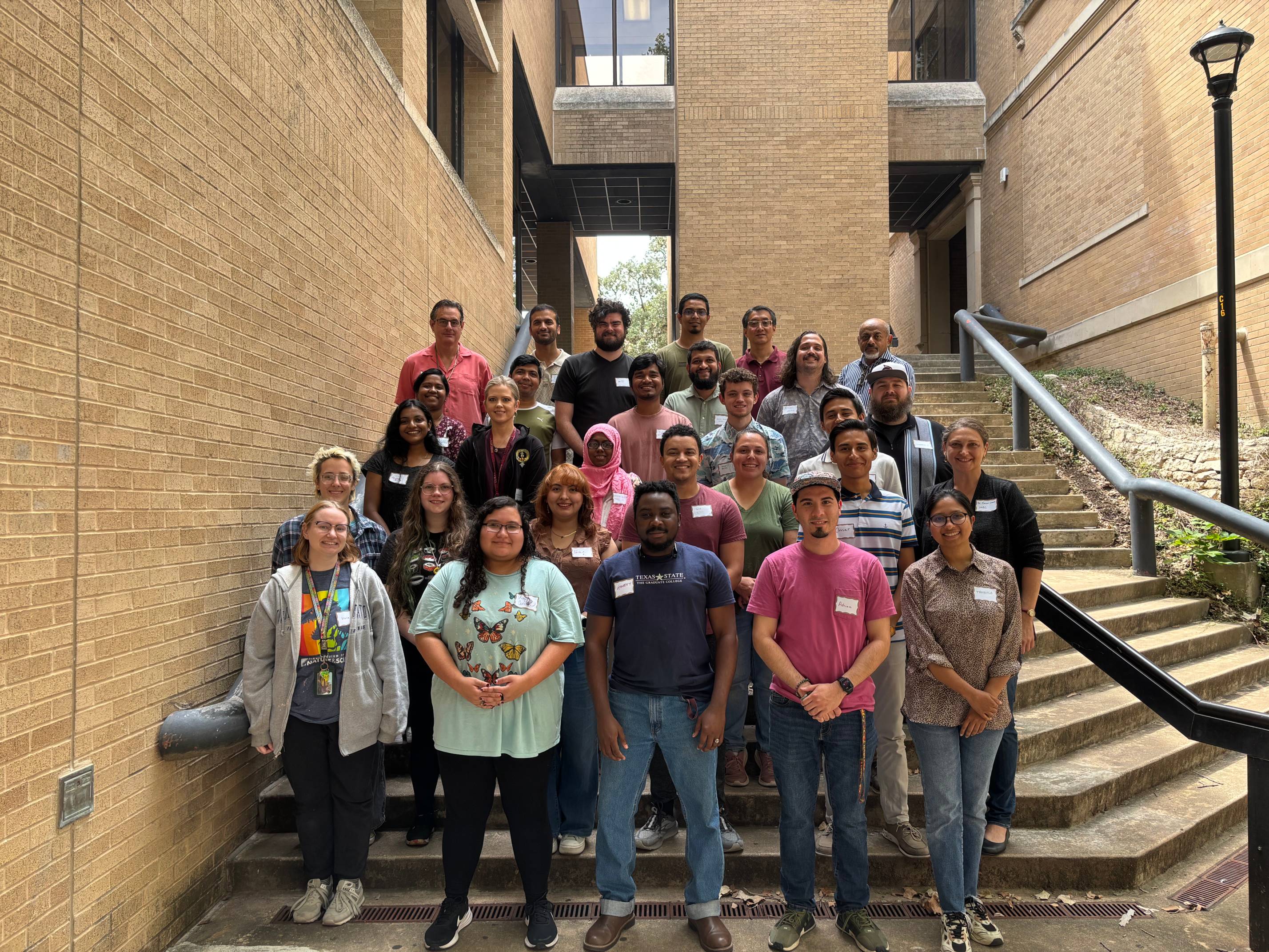  A large group of diverse people, including students and faculty, are gathered on a set of outdoor concrete stairs for a group photo. They are smiling and looking at the camera, standing in rows with a brick building in the background.
