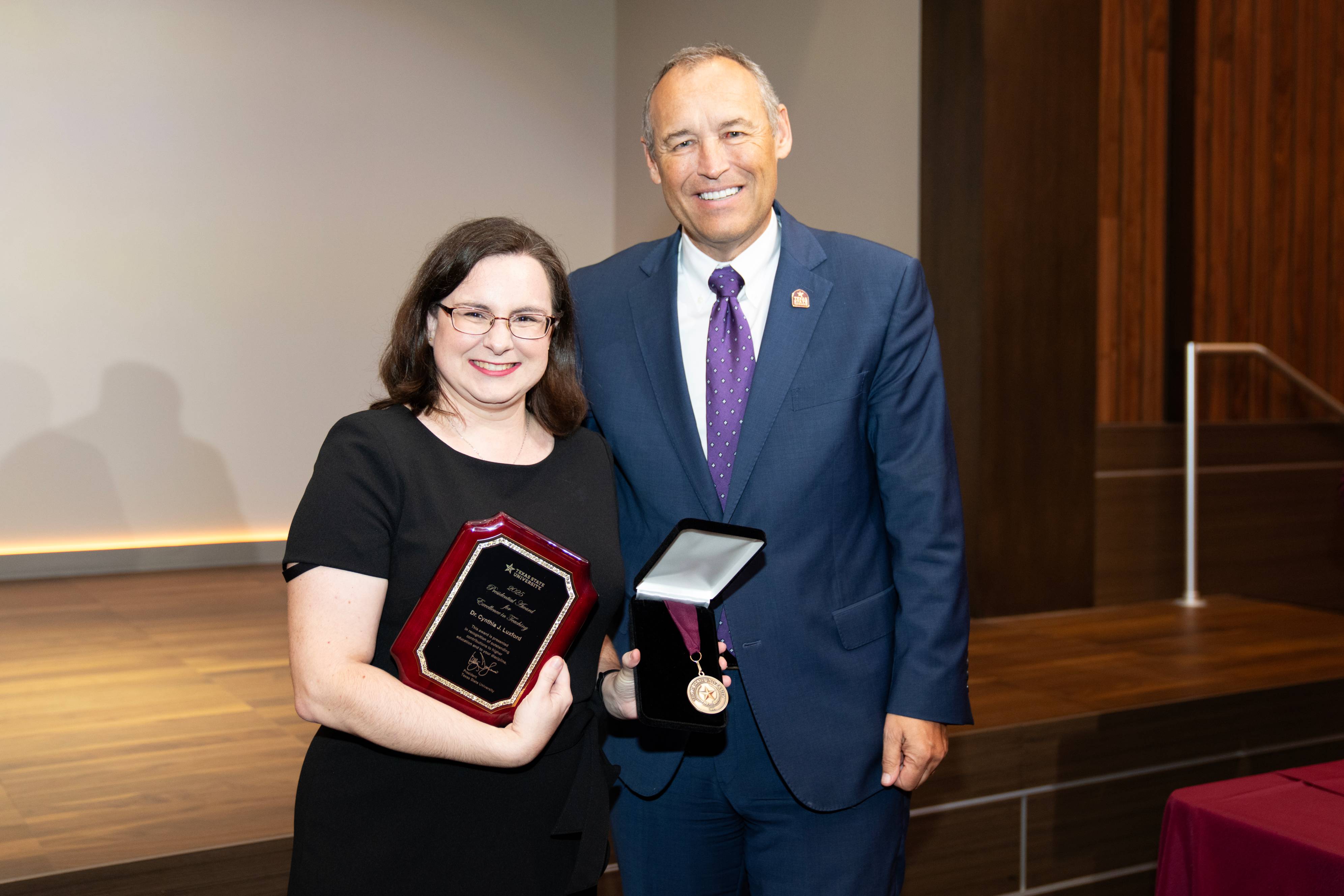  A person in a black dress and glasses holds a plaque with a red cover while standing next to a person in a blue suit, white shirt, and purple tie who holds an open box displaying a medal. Both are smiling and standing on a stage at an awards ceremony.