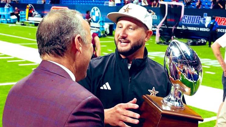 President Kelly Damphousse (left) speaks with Coach GJ Kinne while handing him a trophy.