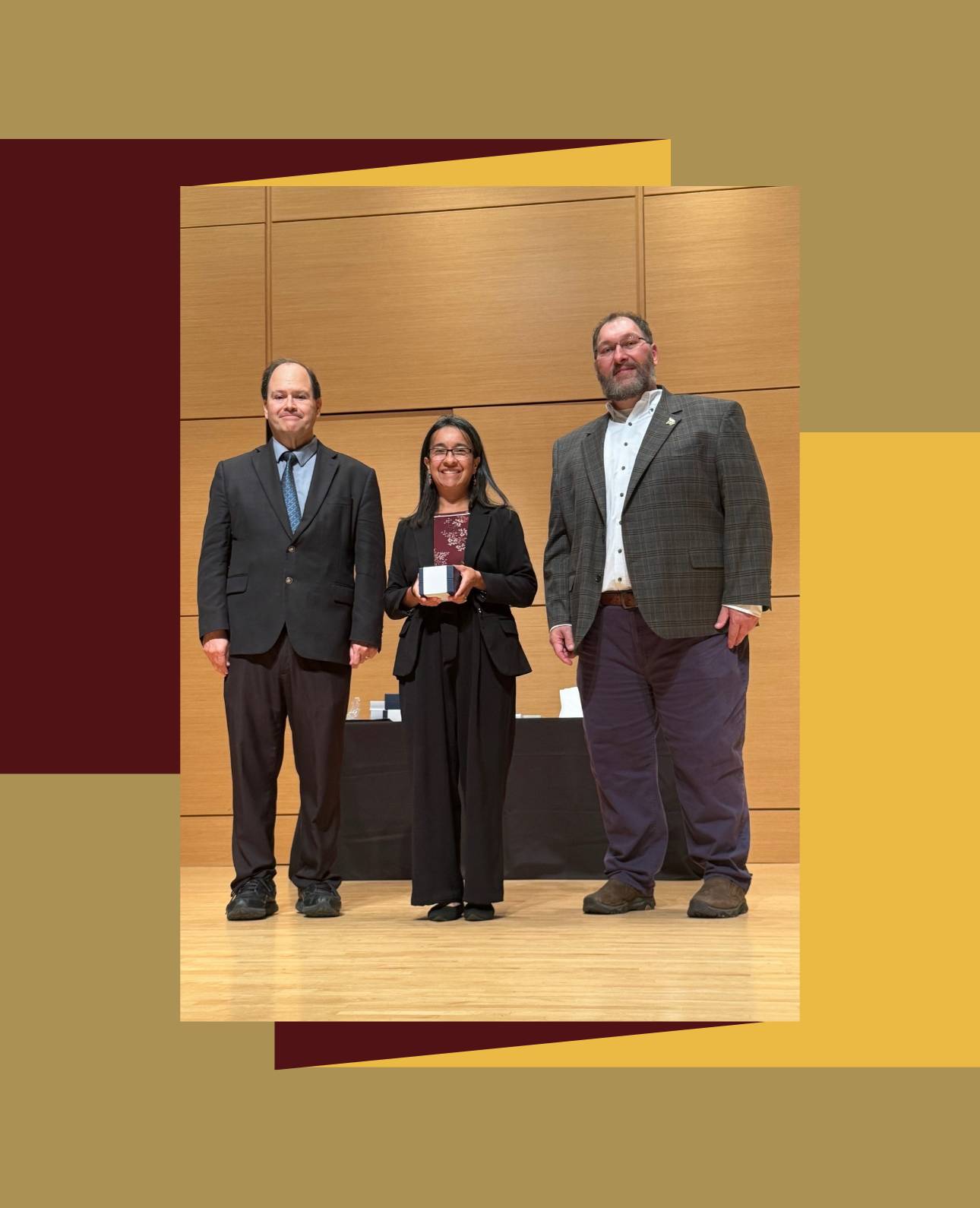  Three people are standing on a stage with a wooden backdrop at an awards ceremony. The person in the center, who is shorter, is wearing a dark suit and is holding a small award. The person on the left is wearing a dark suit and blue tie, and the person on the right is wearing a plaid jacket over a white shirt and dark pants with a beard.