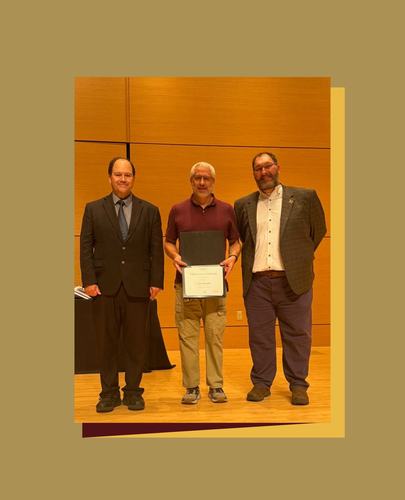  Three people stand on a wooden stage with a wooden backdrop at an awards ceremony. The person in the center is wearing a maroon polo shirt and khaki cargo pants, holding a certificate, and is flanked by two people in suits, one on the left in a dark suit and tie, and one on the right in a plaid jacket and dark pants. 
