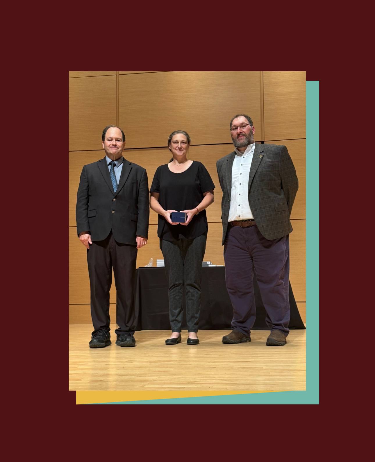 Three people are standing on a wooden stage with a wooden backdrop at an awards ceremony. The person in the center is wearing a black top and dark pants, holding a small award. They are flanked by two people in suits: one on the left in a dark suit and tie, and one on the right in a plaid jacket and dark pants. 