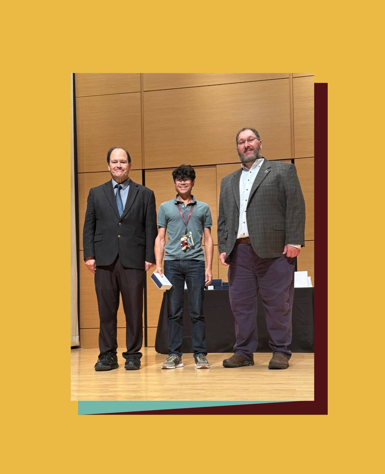 Three people stand on a wooden stage with a wooden backdrop at an awards ceremony. The person in the center is wearing a polo shirt, jeans, and a lanyard, holding a small award. They are flanked by a person in a dark suit and tie on the left, and a person in a plaid jacket and dark pants on the right.