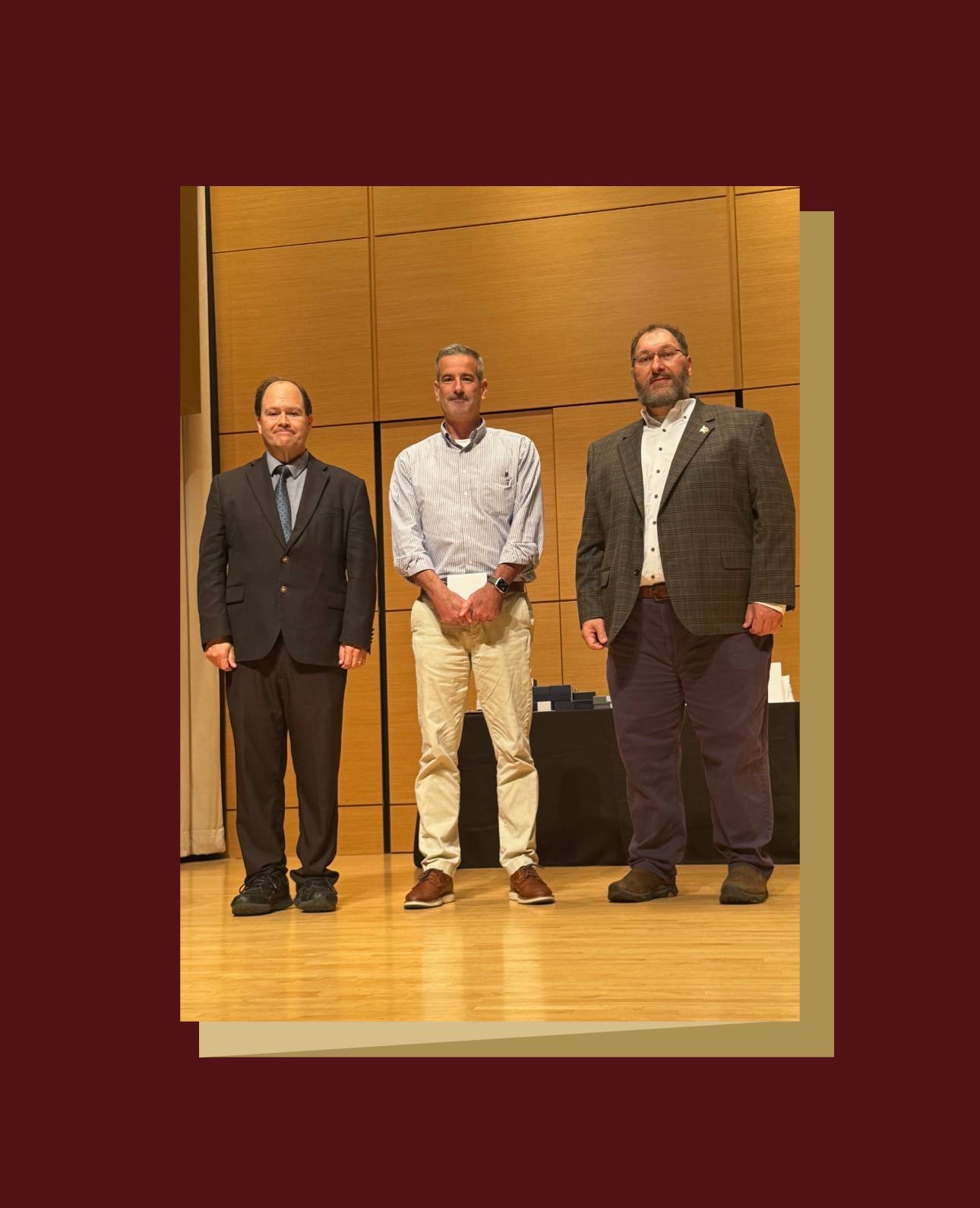 Three people stand on a wooden stage with a wooden backdrop at an awards ceremony. The person in the center is wearing a light-colored striped shirt and khaki pants. They are flanked by a person in a dark suit and tie on the left, and a person in a plaid jacket and dark pants on the right. 