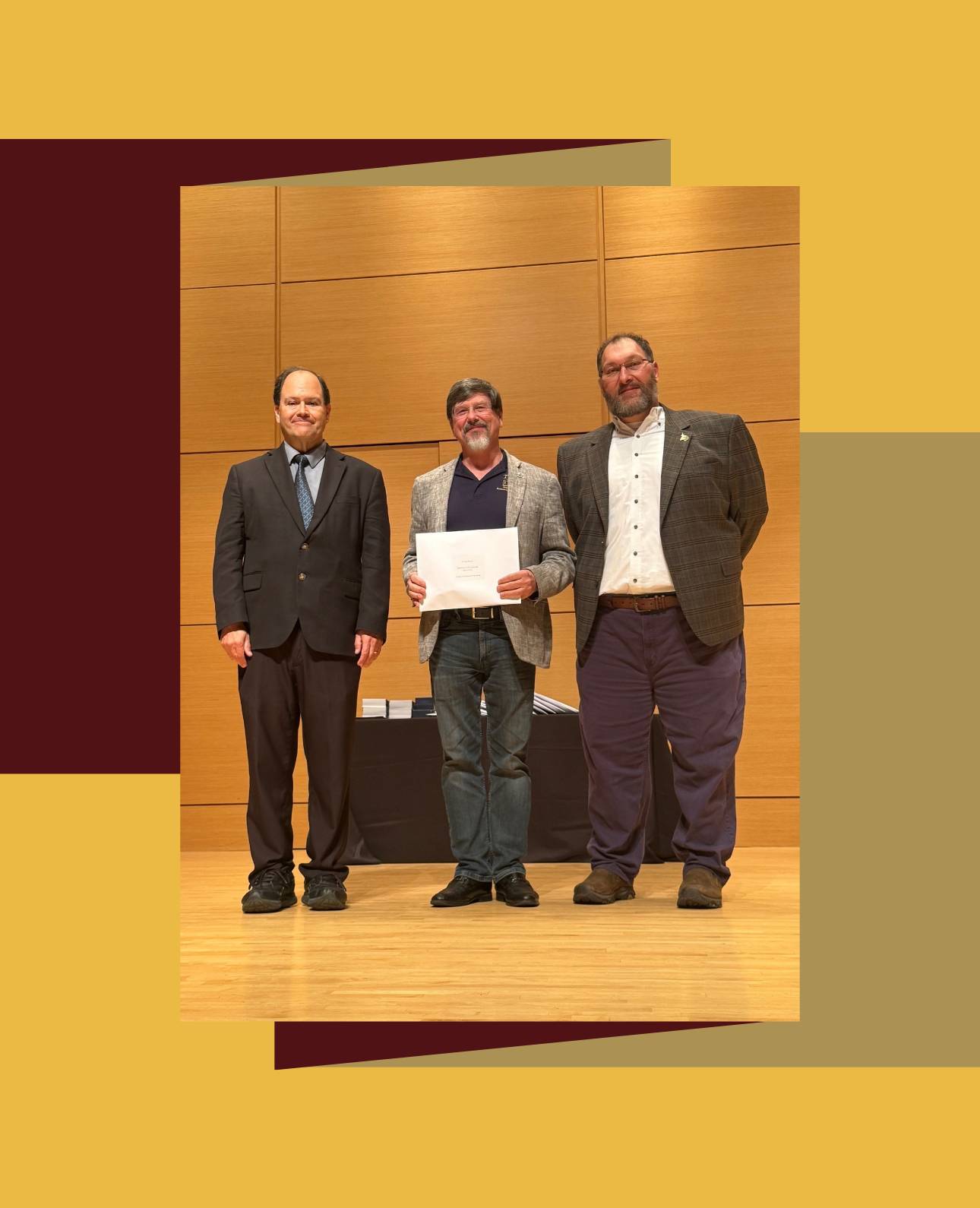  Three people stand on a wooden stage with a wooden backdrop at an awards ceremony. The person in the center is wearing a blazer, a polo shirt, and jeans, holding a white certificate. They are flanked by a person in a dark suit and tie on the left, and a person in a plaid jacket and dark pants on the right. 