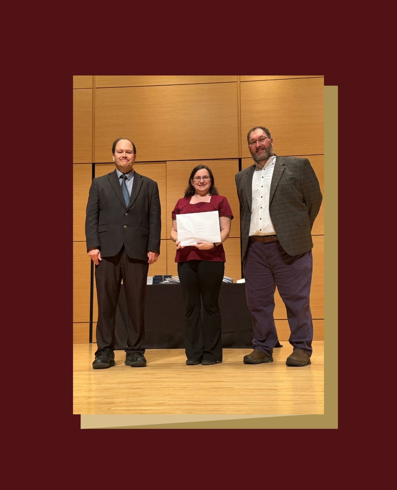  Three people stand on a wooden stage with a wooden backdrop at an awards ceremony. The person in the center is wearing a maroon top and black pants, holding a white certificate. They are flanked by a person in a dark suit and tie on the left, and a person in a plaid jacket and dark pants on the right.