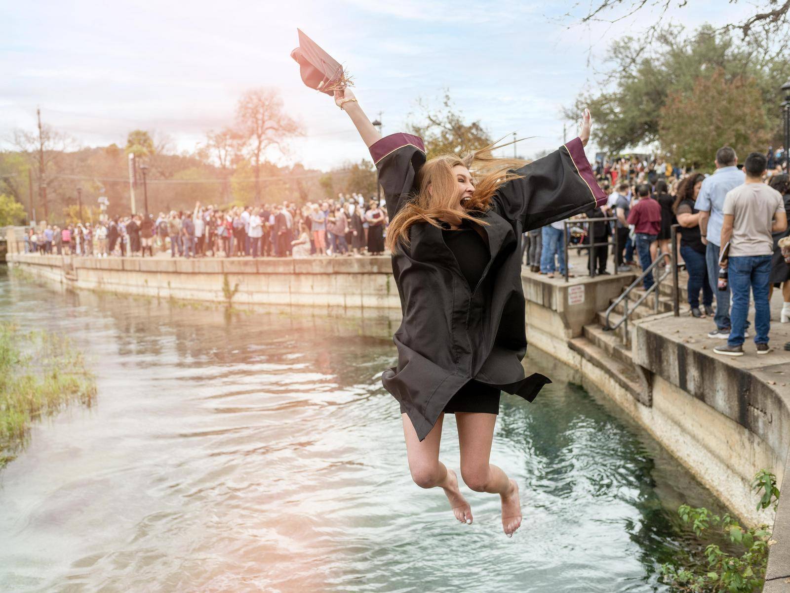 Young woman in her commencement robe joyfully leaps into the San Marcos River after her graduation ceremony.