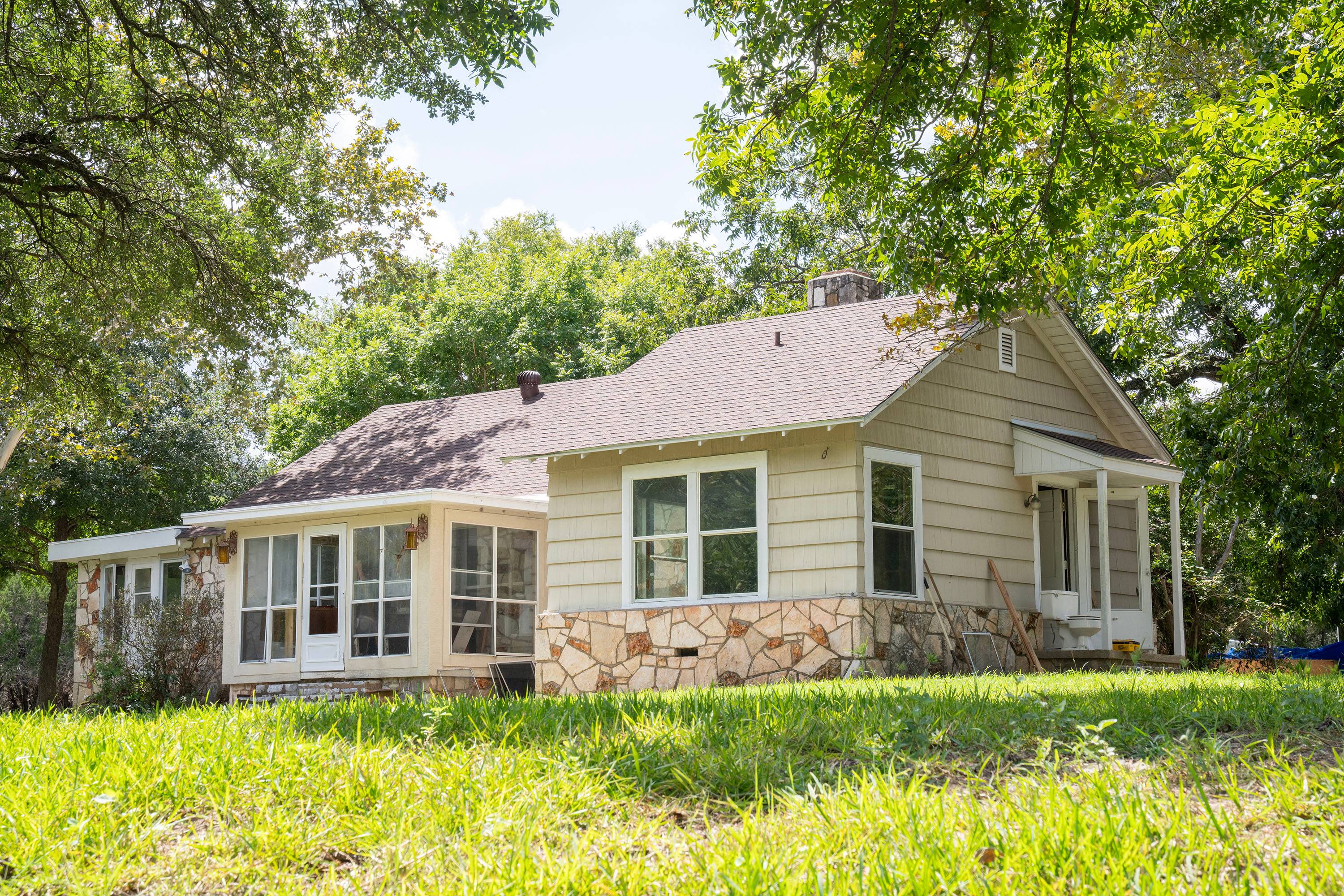 A single story house with cream siding sits on a hill with green grass and trees.