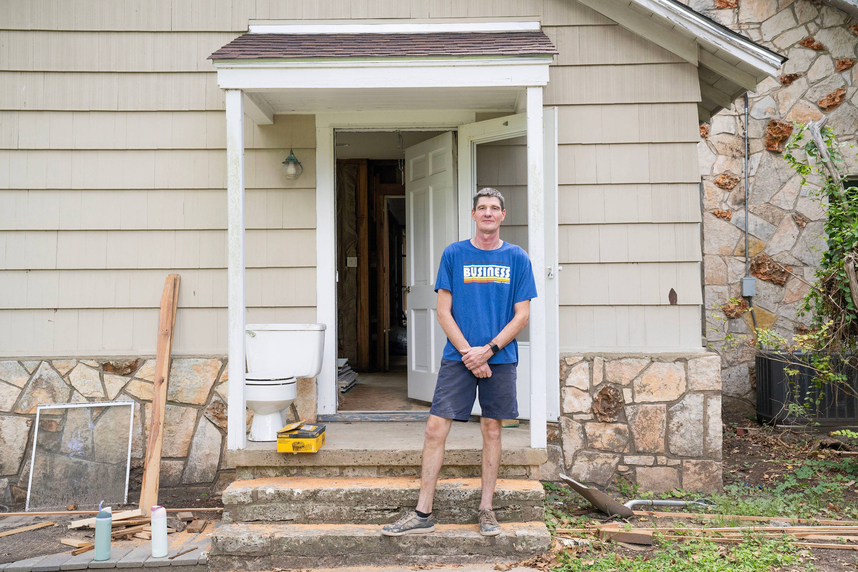 A man in a blue shirt stands just outside of a house with cream colored siding.
