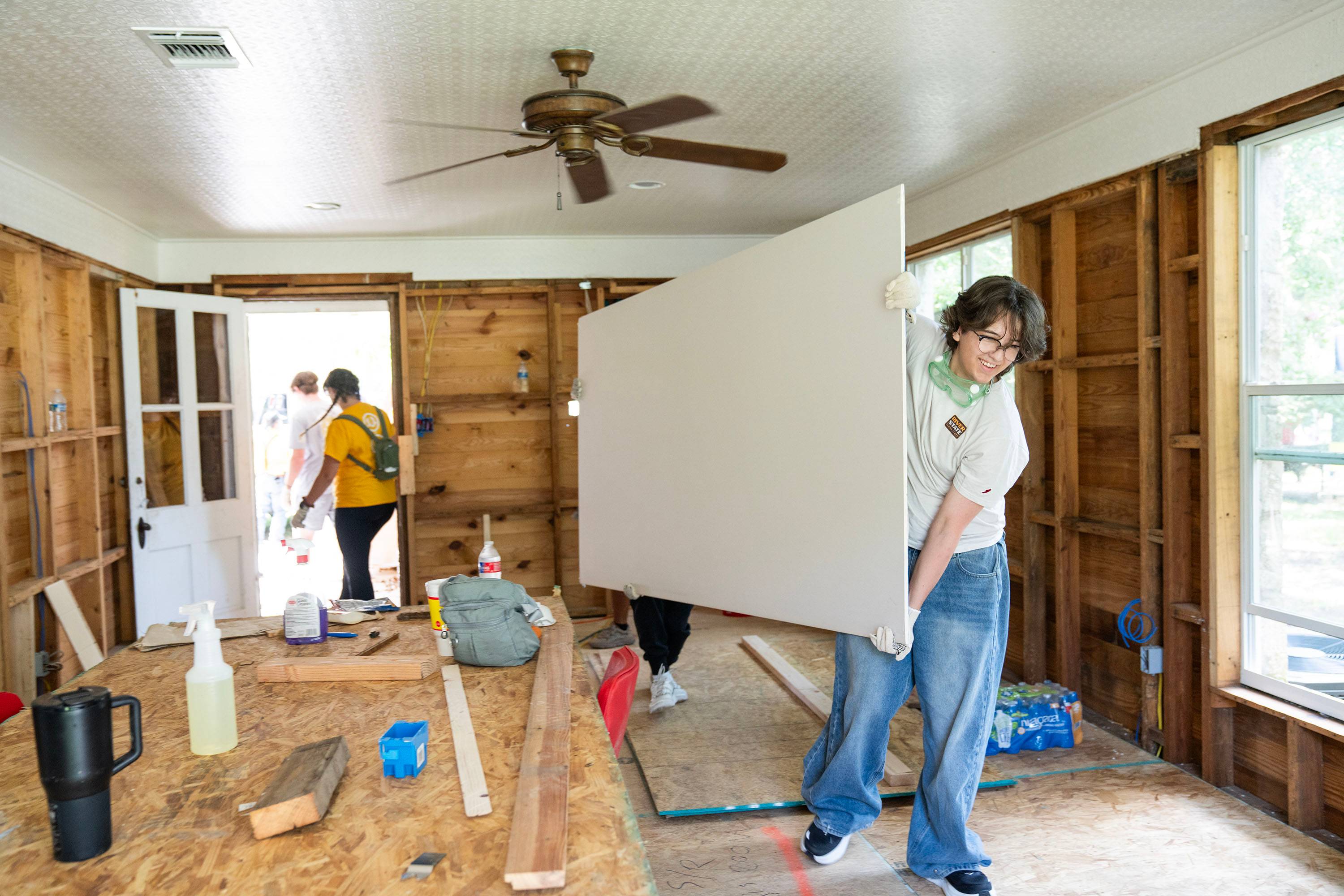 Two students carry a piece of sheet rock into a house.