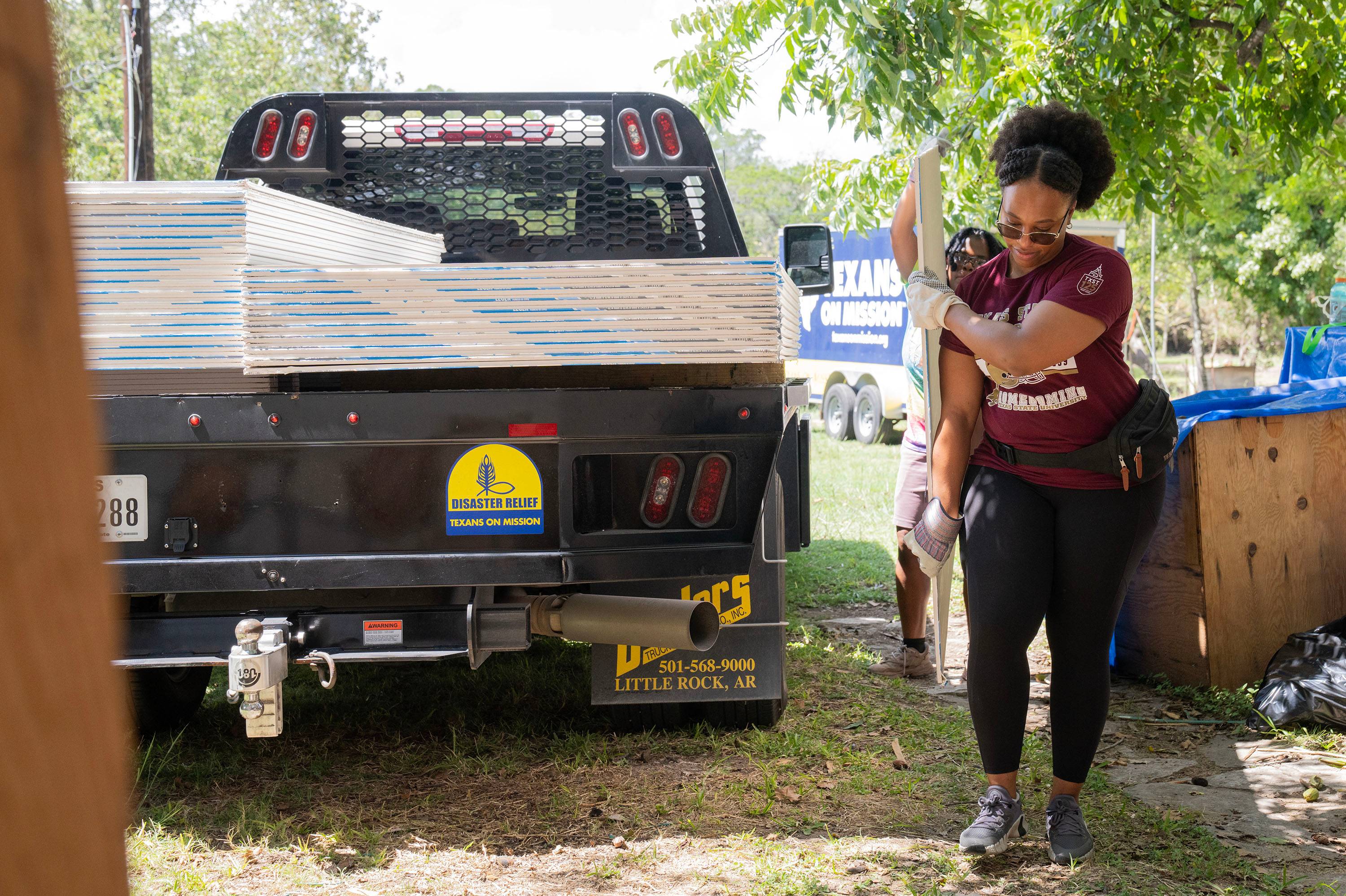 Two people carry a piece of sheetrock off of a truck loaded with several sheets.