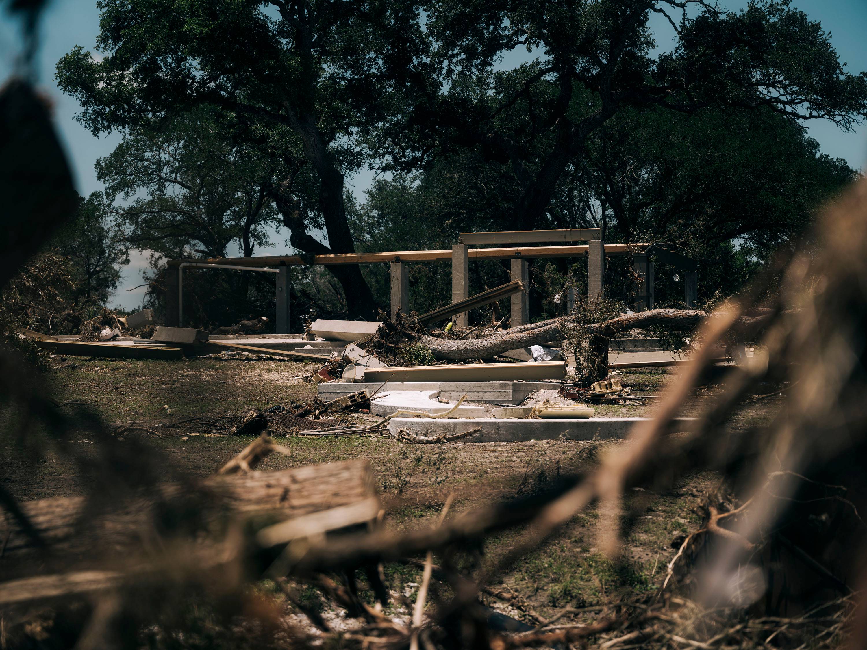 The remains of a house, just now several concrete pillars, stands surrounded by trees and debris. other debrei. 