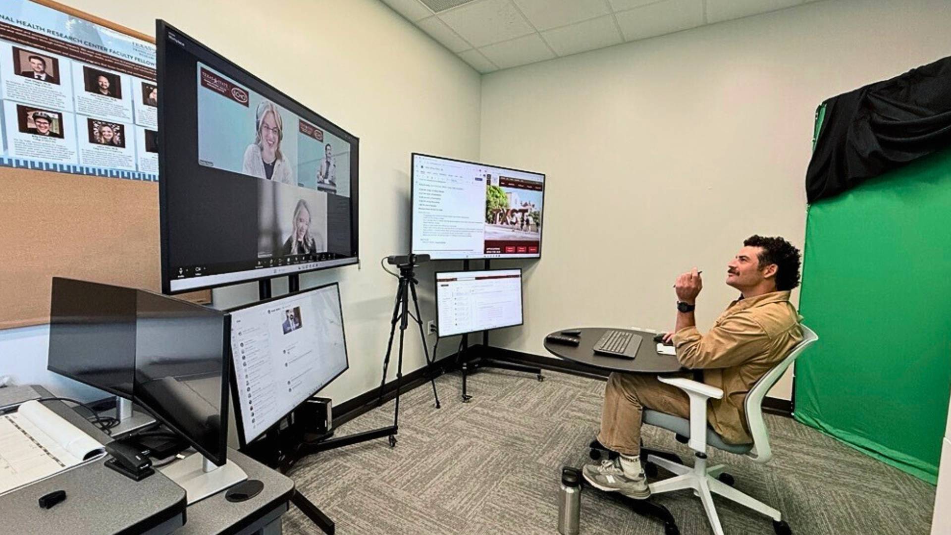 Cody Gould sits a a table in a desk chair facing four large monitors where MHCC guest speakers can be seen on Zoom.