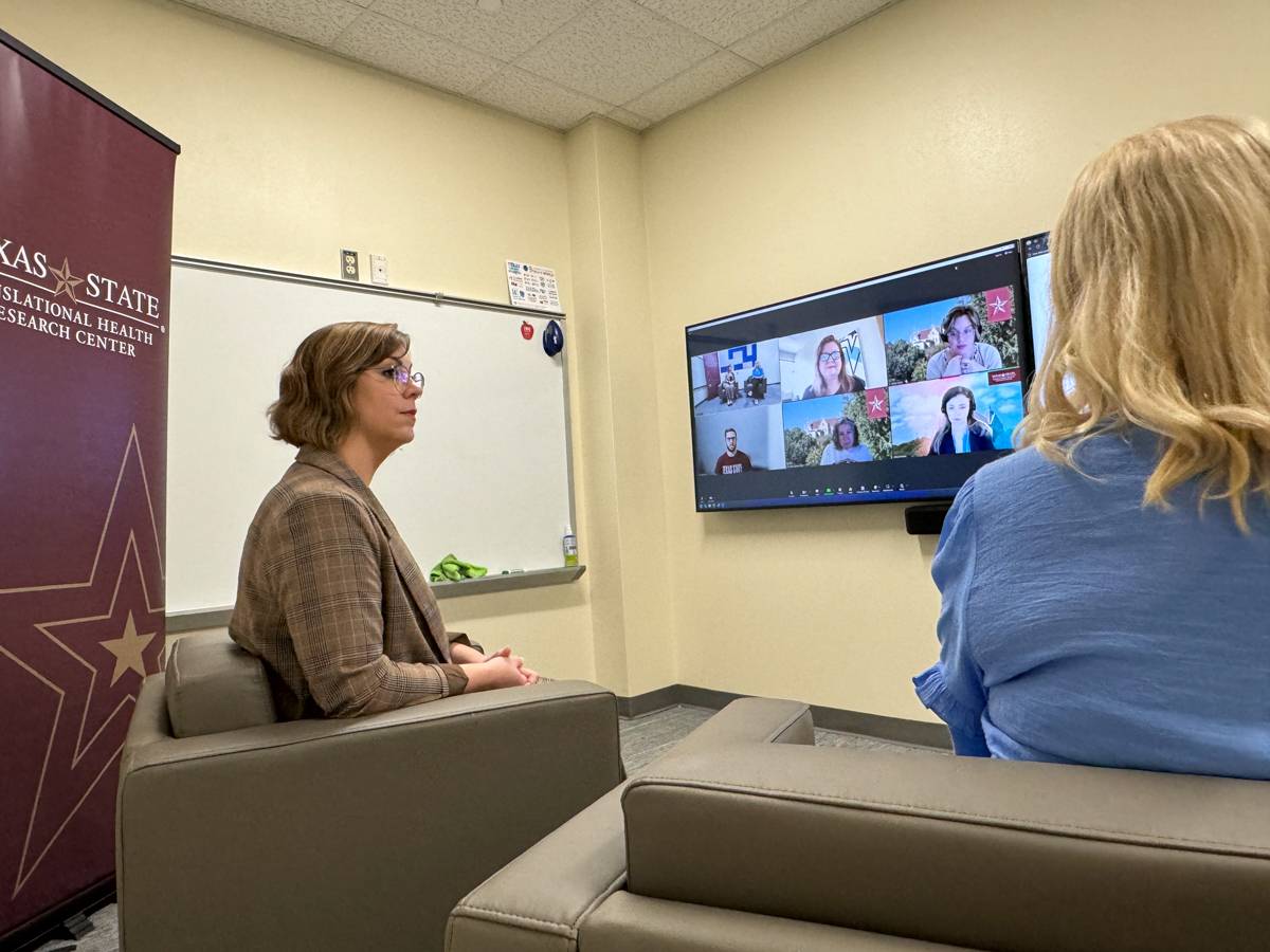 Jessica Schneider and Dr. Melinda Villagran sit in chairs facing large monitors. Zoom participants can be seen on the screen.