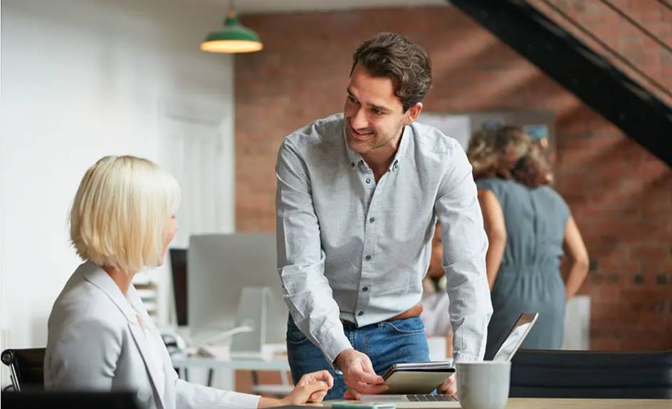 a man standing next to a desk where a woman is seated