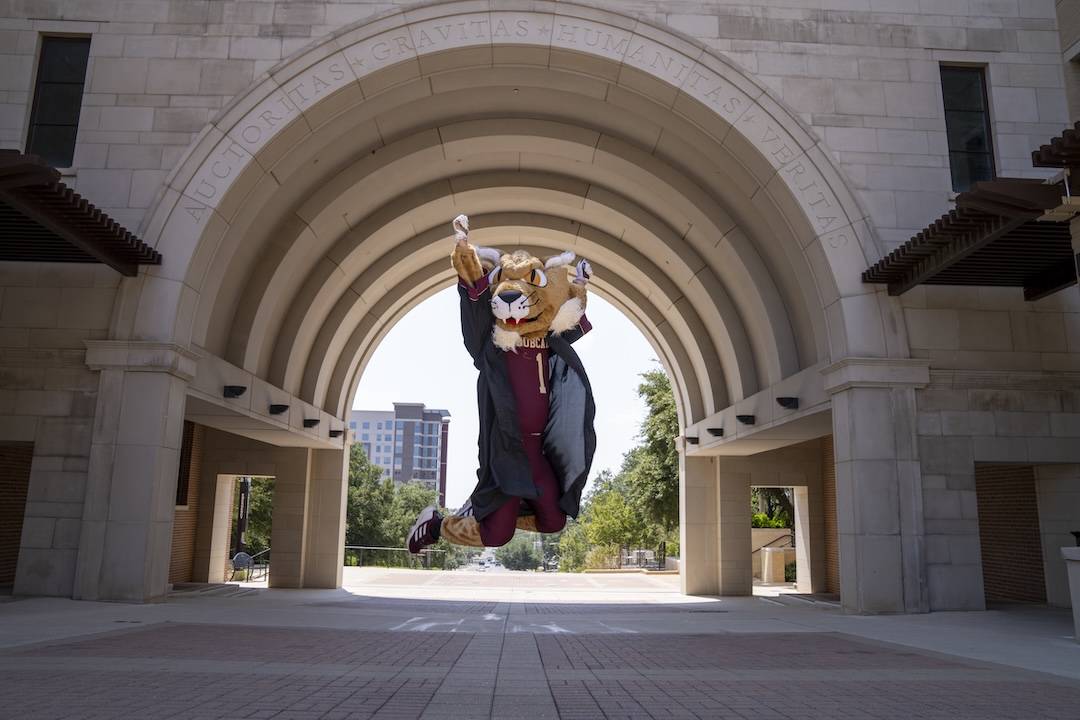 Boko jumping in grad gown in front of UAC arch. 