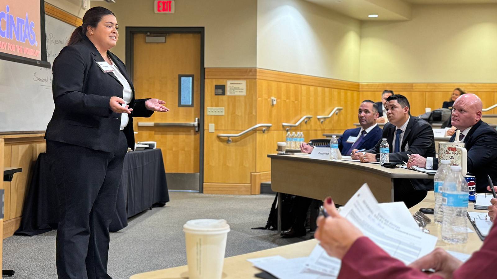 professionally dressed student standing in front of classroom delivering sales pitch to group of individuals