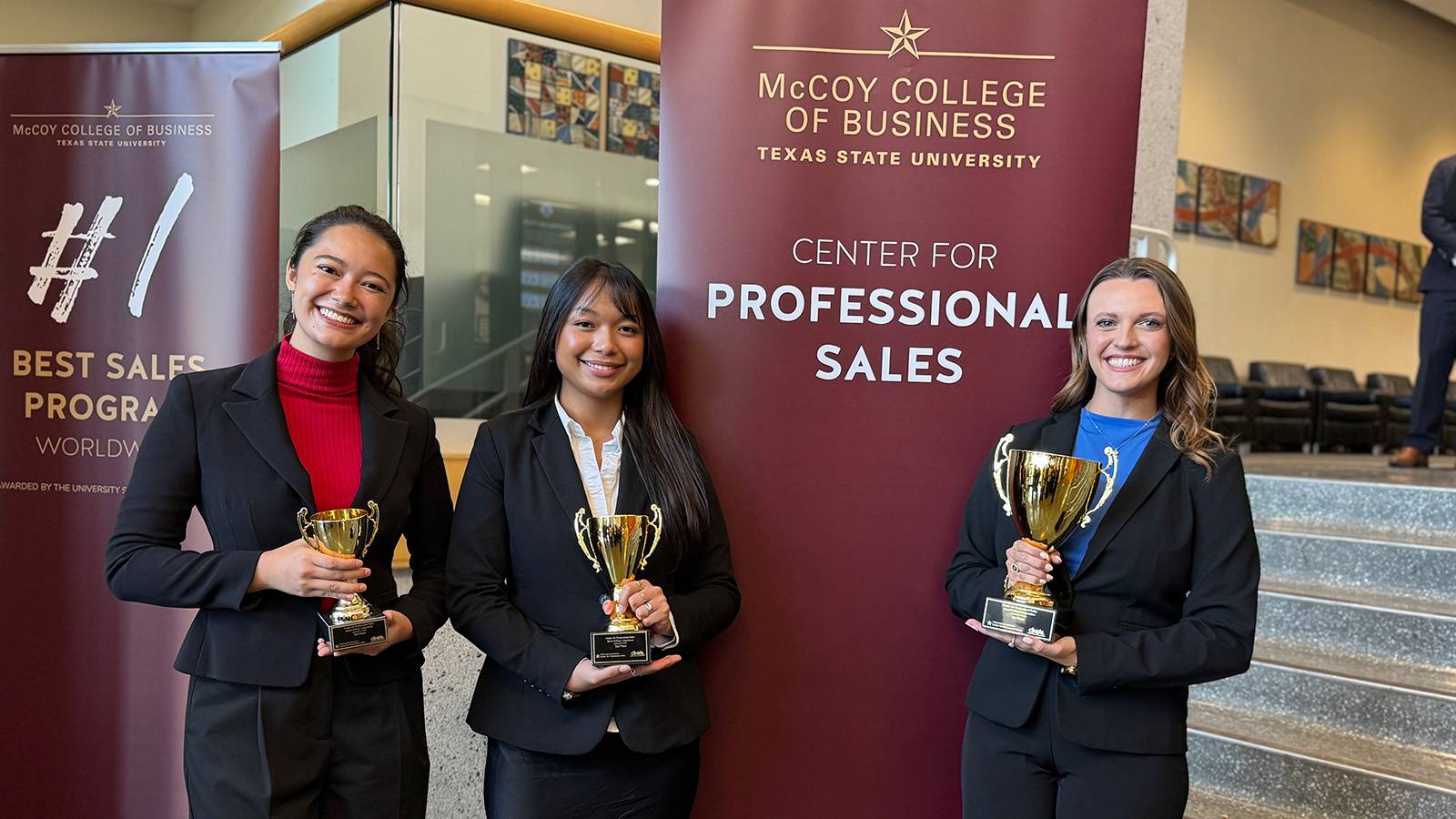 three female professionally dressed students holding trophies and smiling for a photo