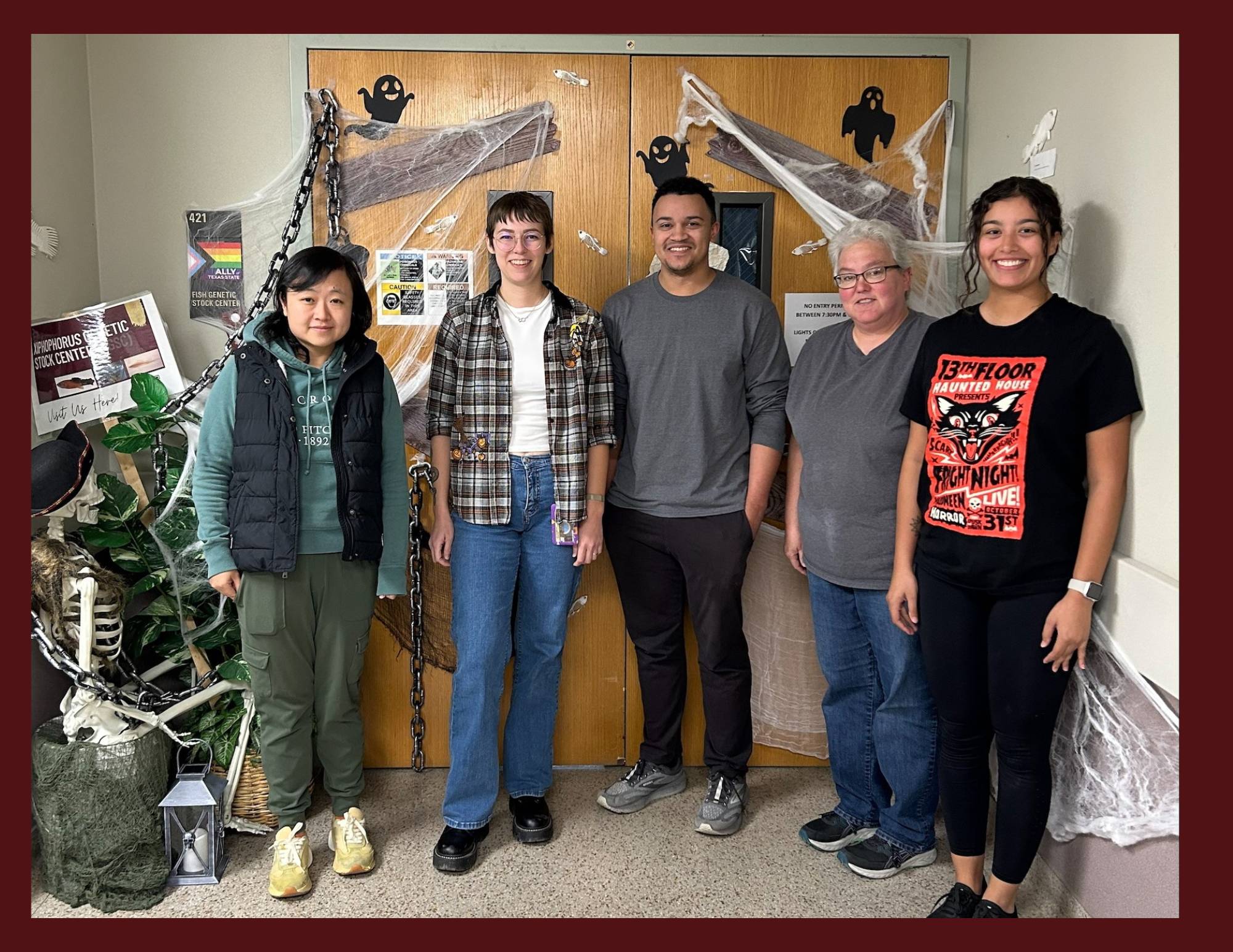 A diverse group of five people stands in a hallway in front of a double door decorated for Halloween with chains, cobwebs, and paper ghosts.