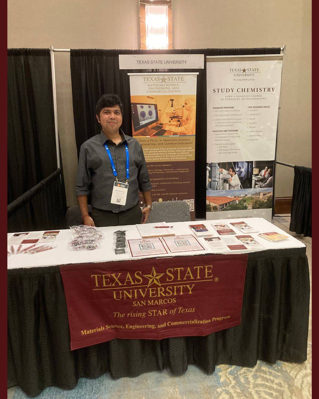 A person stands behind an informational table for the Texas State University Materials Science, Engineering, and Commercialization program at a professional event. The table features a maroon banner, brochures, and two large vertical banners detailing graduate programs and research in Materials Science and Chemistry.
