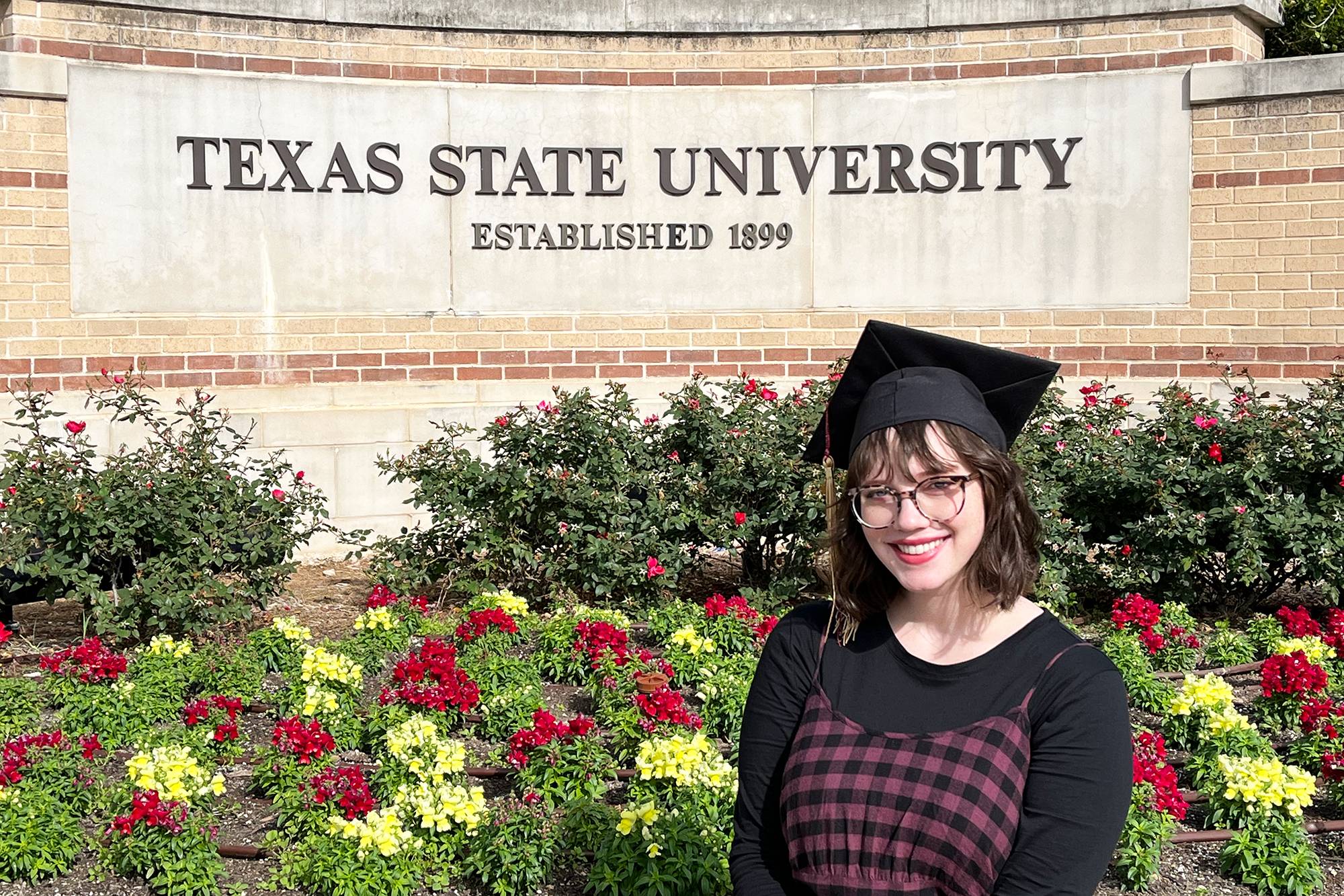 Abby Parker wearing a graduation cap in front of a Texas State University sign