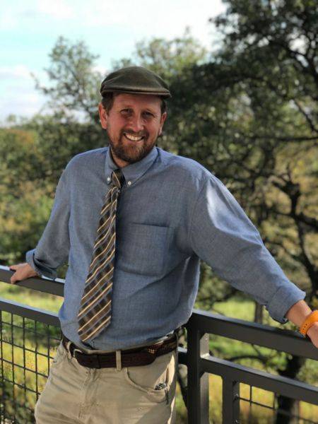 man wearing a button up shirt, tie and hat on his head leans against a fence smiling at camera