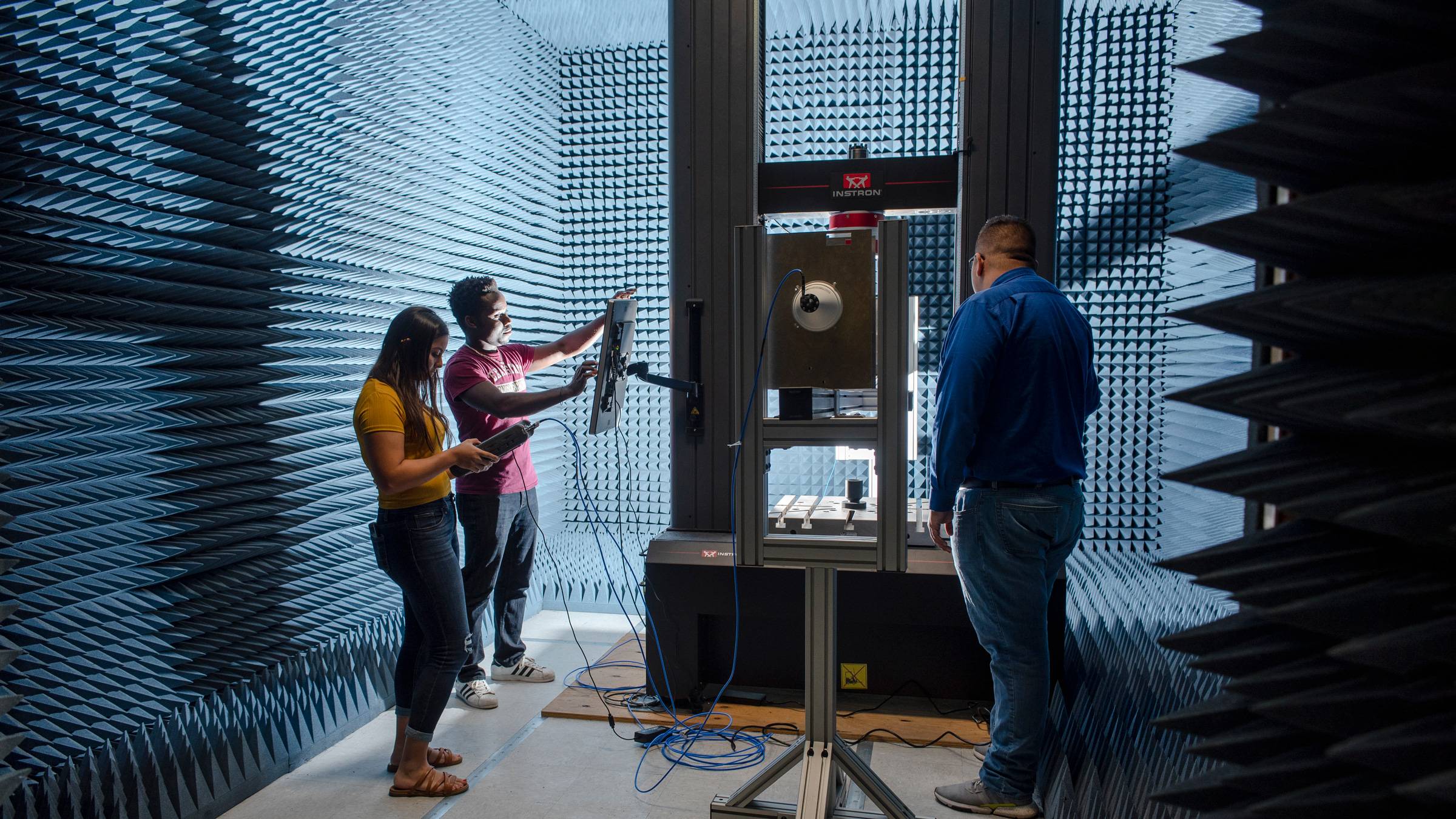 A professor looks on as two students input data in a blue, padded sound lab.