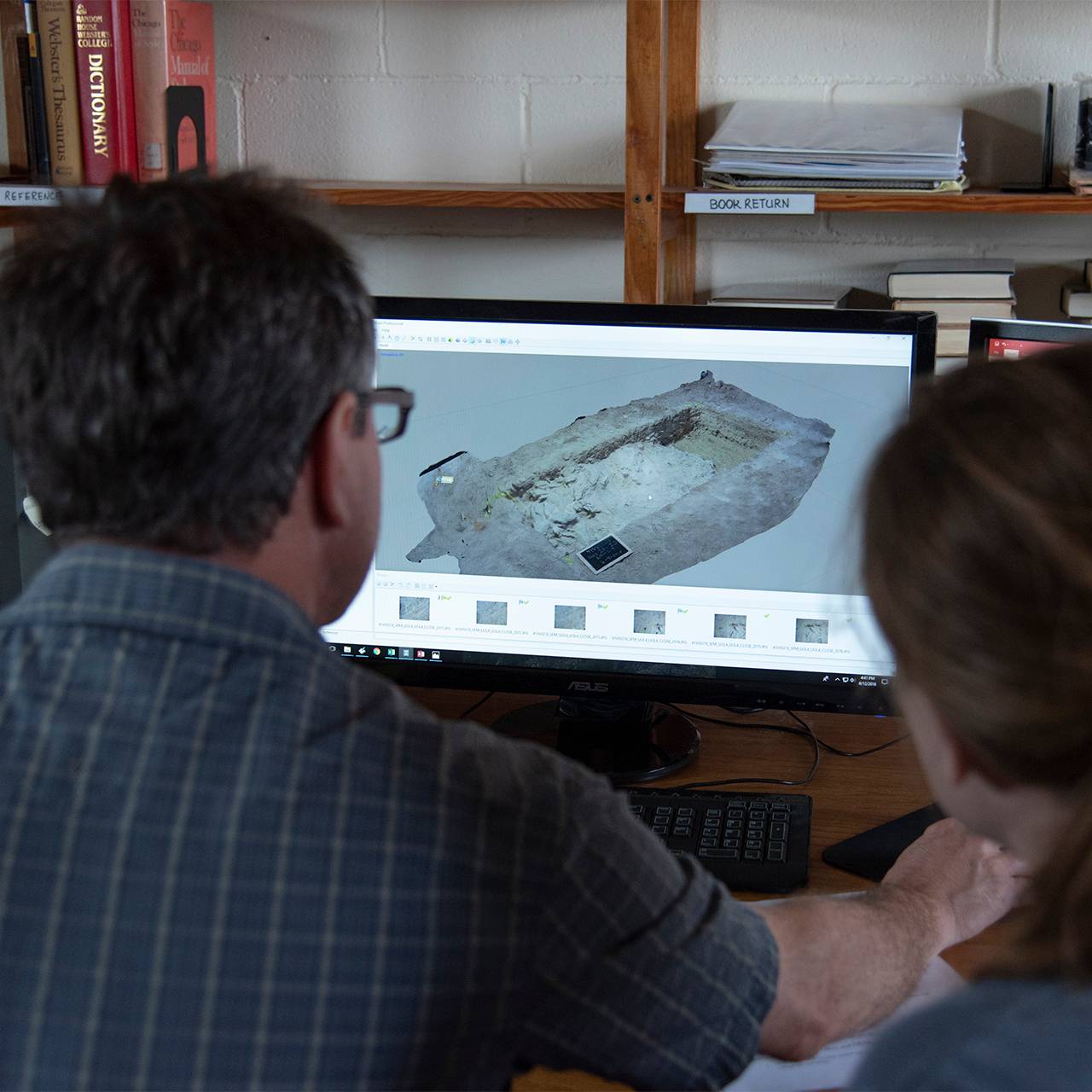 A man and woman huddle around a screen displaying photos of an archaeological dig.