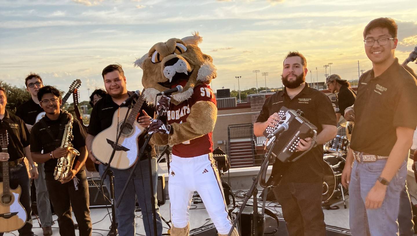 A bobcat mascot stands outside with a band, members wearing black polos and jeans. 