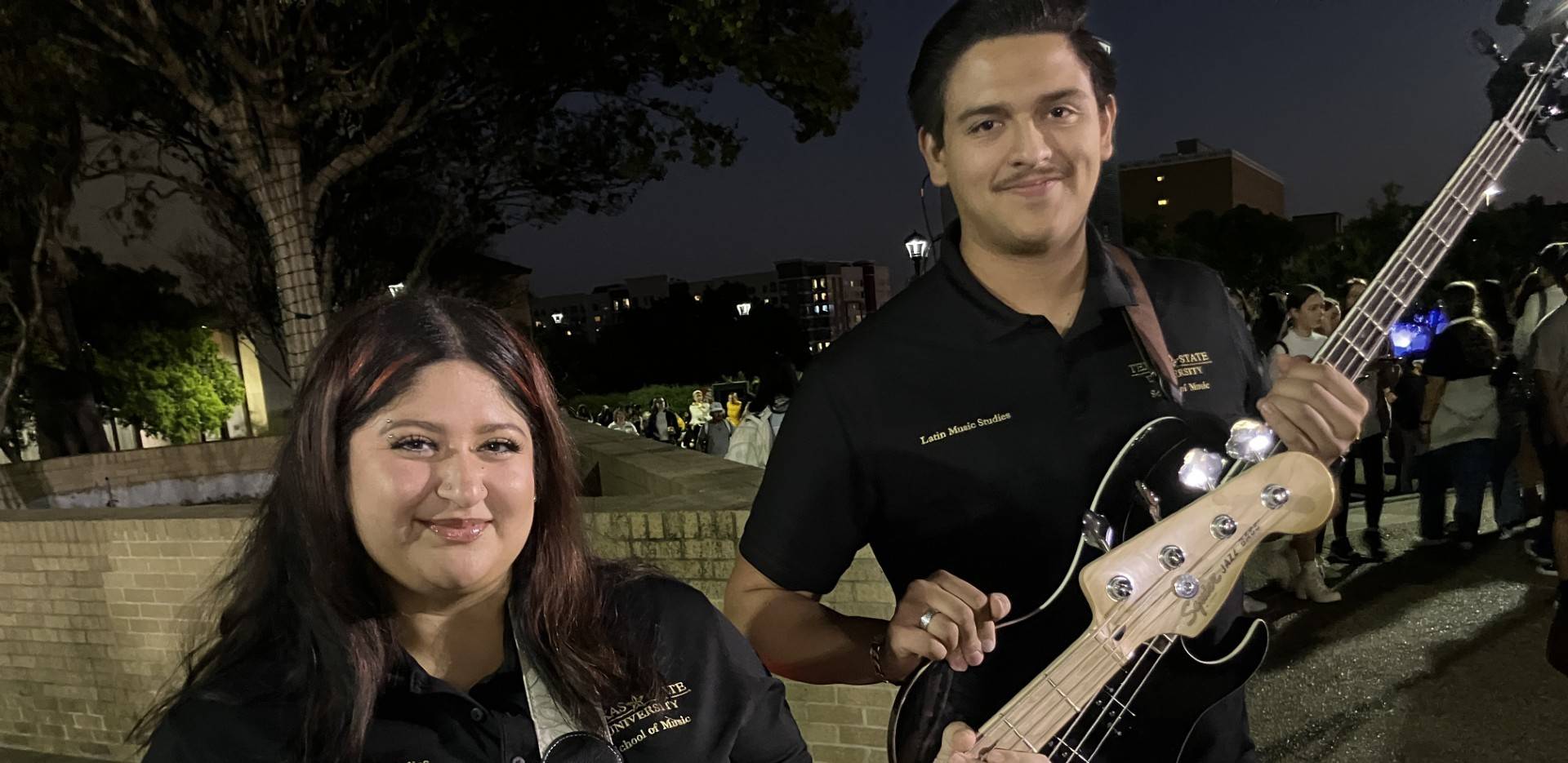 Two students wearing black polos are holding guitars. It is nighttime and outside.