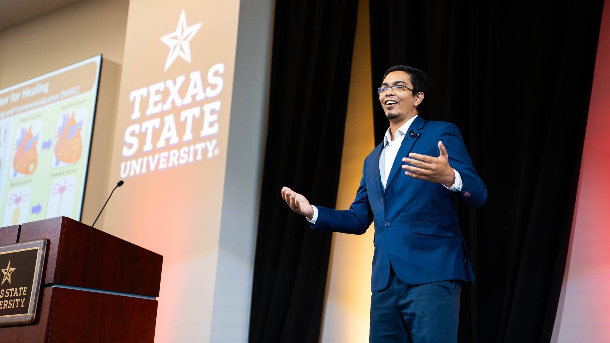  A person in a blue suit and glasses is giving a presentation, standing next to a wooden podium with a microphone. The background features a large screen displaying the Texas State University logo and an image of a heart related to the presentation topic.