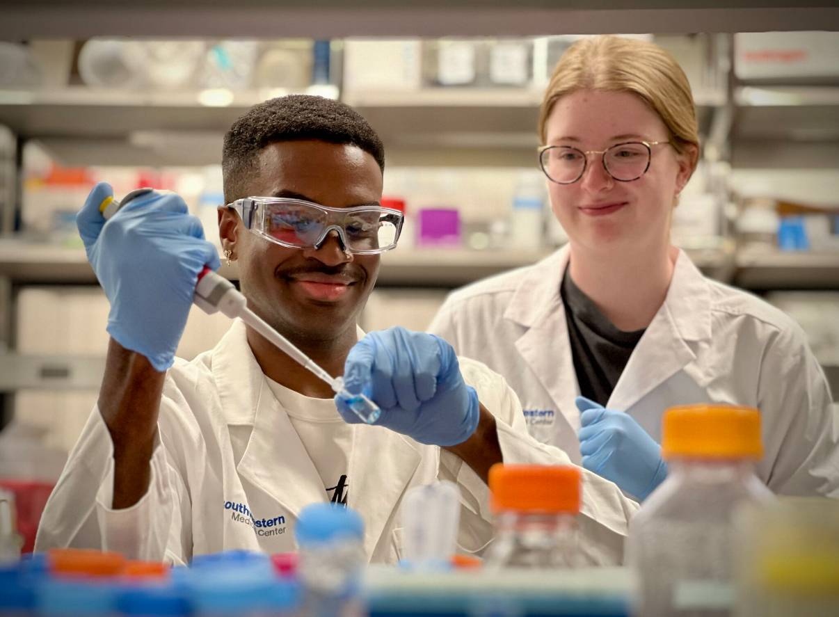  Two people in lab coats and gloves are working in a laboratory. The person in the foreground, wearing safety goggles, is using a pipette to transfer liquid into a small vial, while the person behind them smiles.