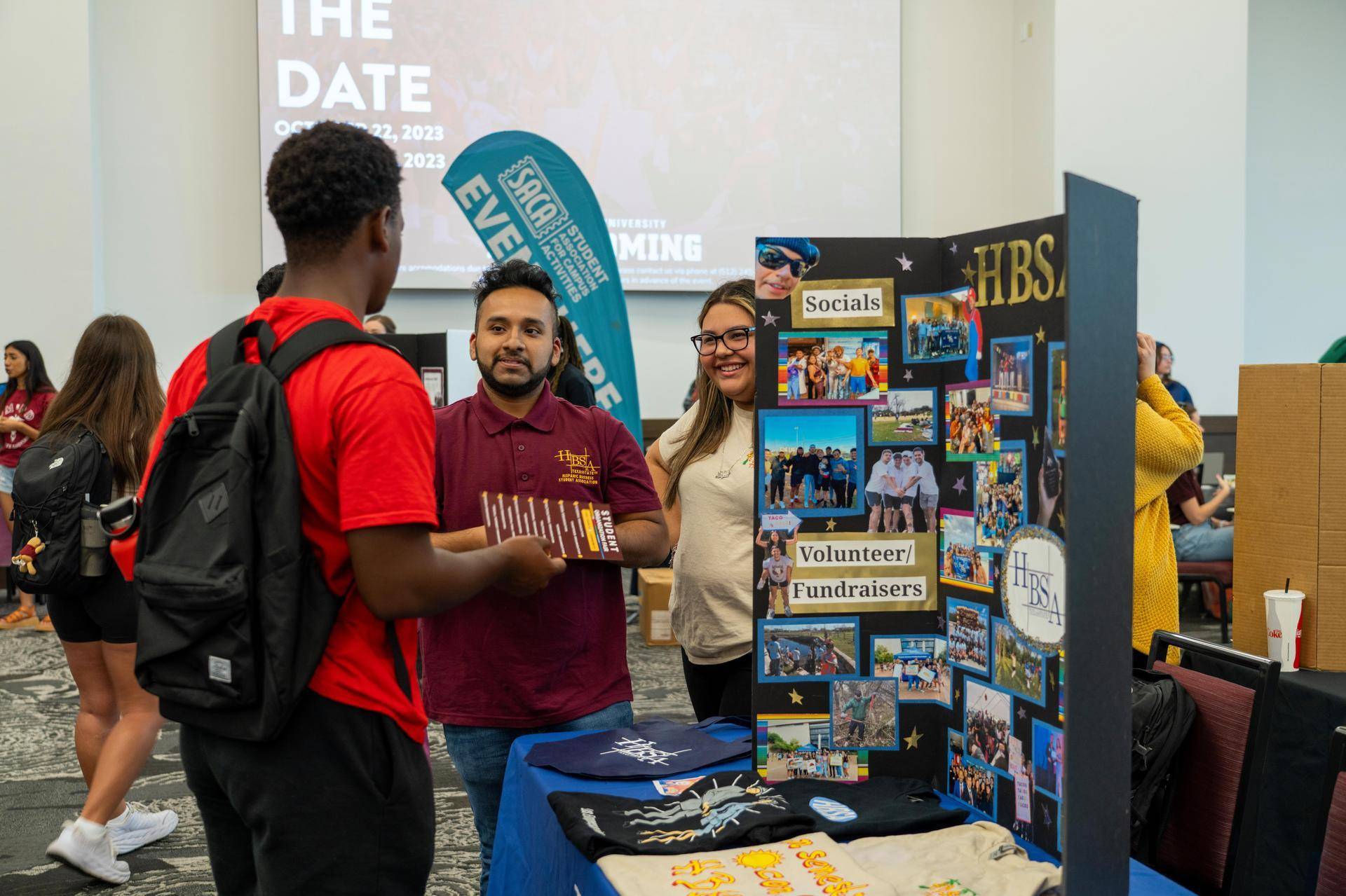 A student stops to look at a display and chat with representatives of a student organization in a crowded hall. 