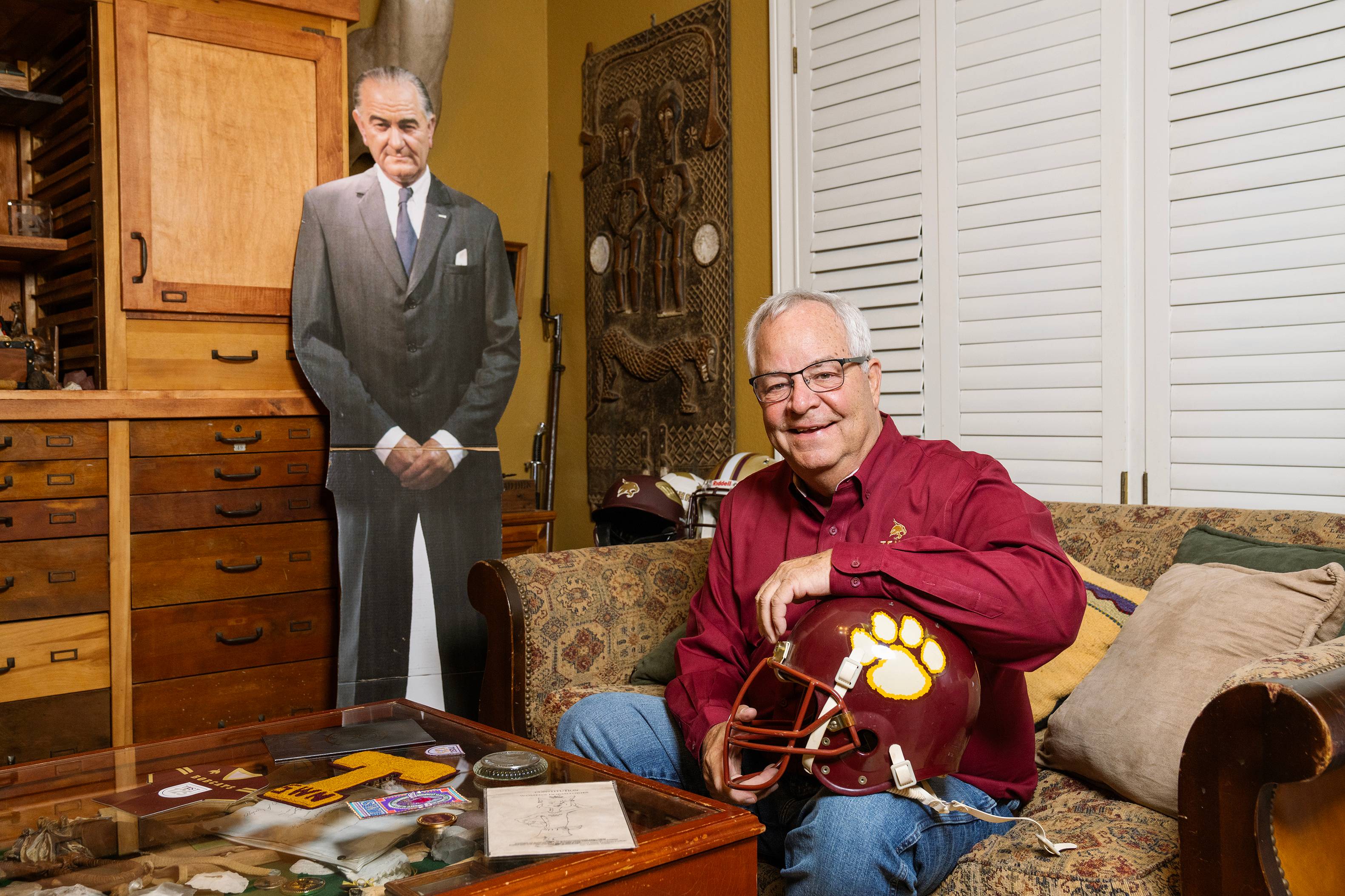 Tom Madden sits on a couch while resting a maroon Bobcats football helmet on his knee. A cardboard cut out of LBJ stands behind him.