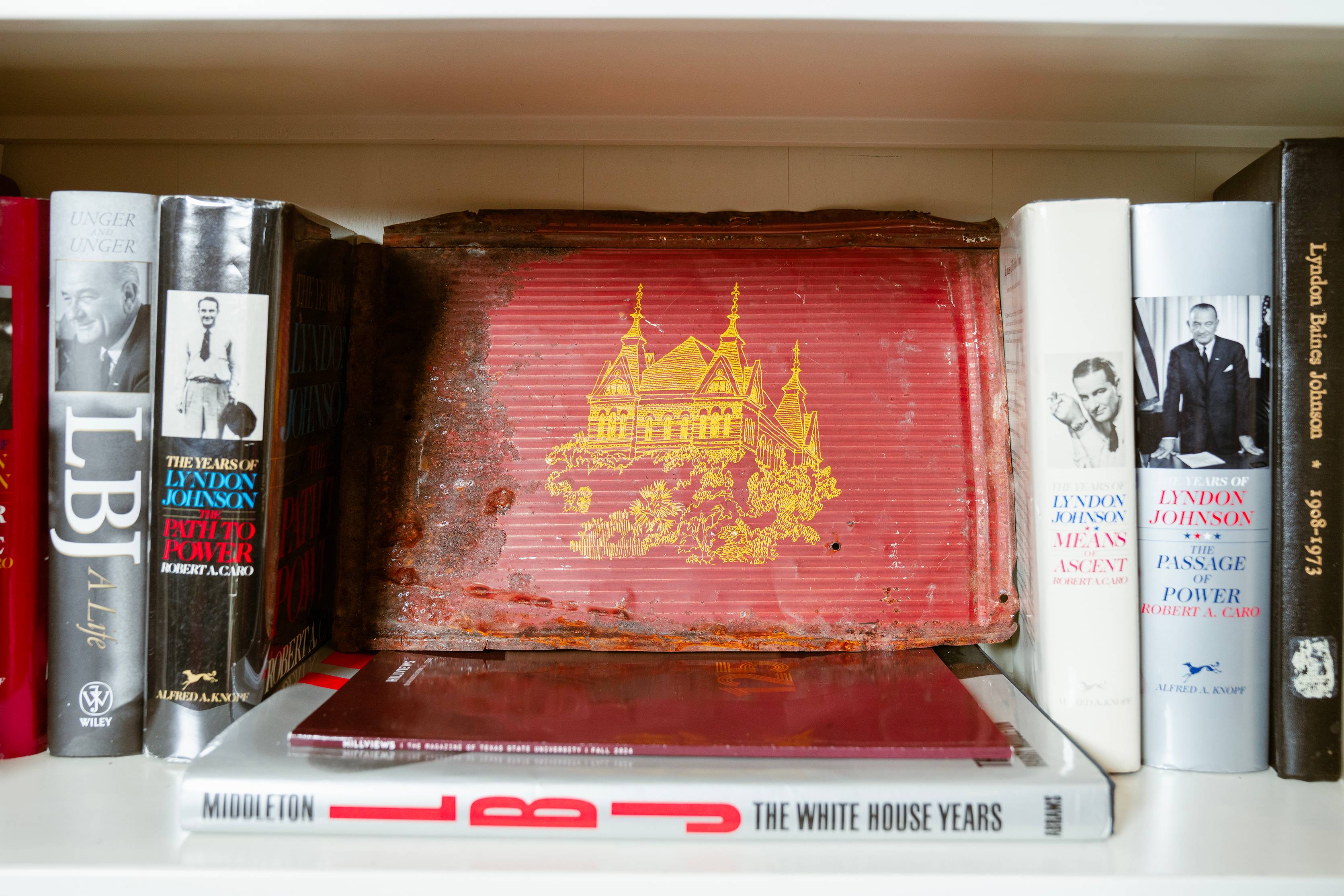 a metallic roof single with a gold painting of a building sits between books about LBJ on a bookshelf