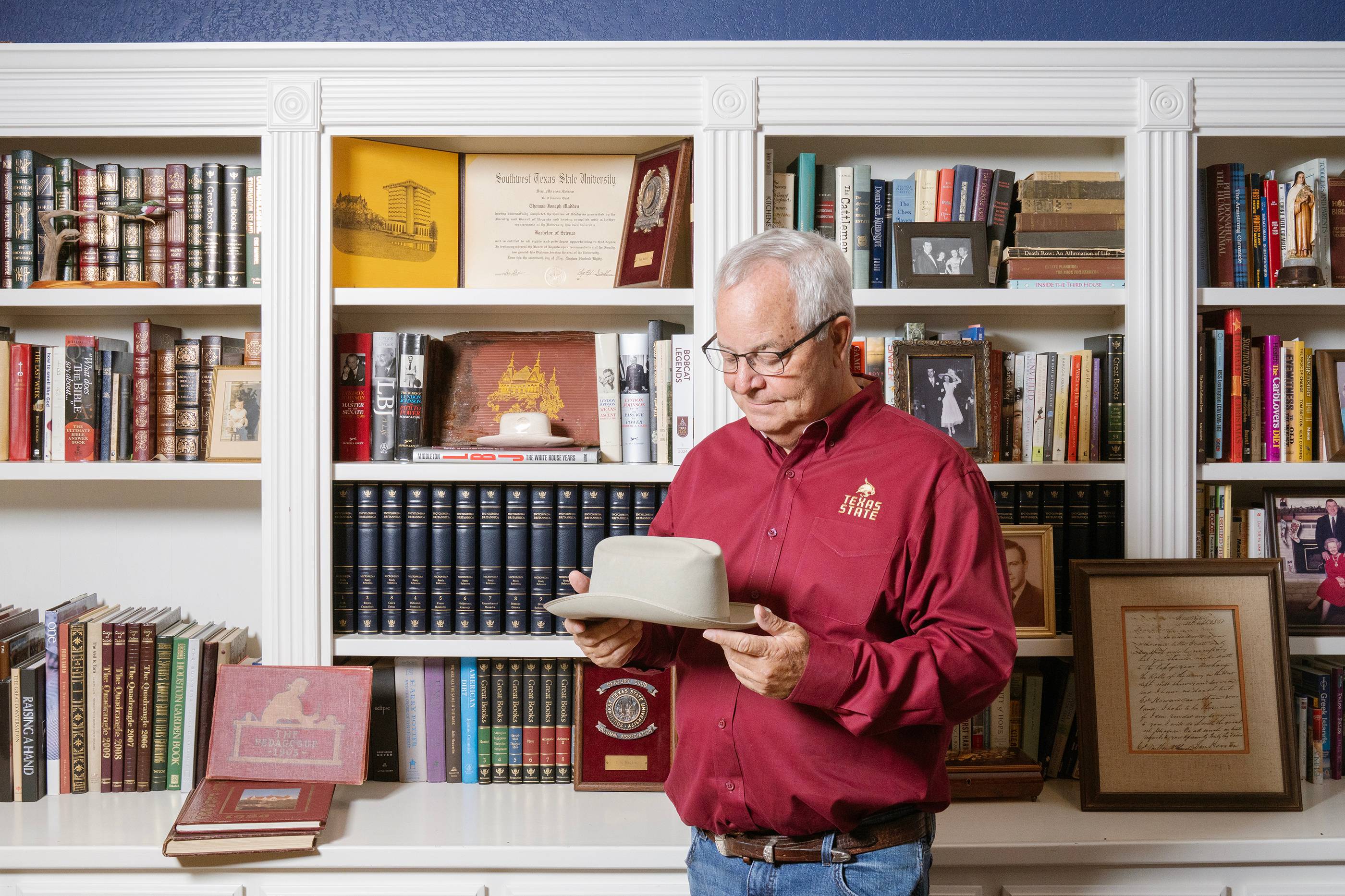 man in maroon button down shirt standing in front of a white bookshelf and looks at Stetson hat he is holding in his hands. 