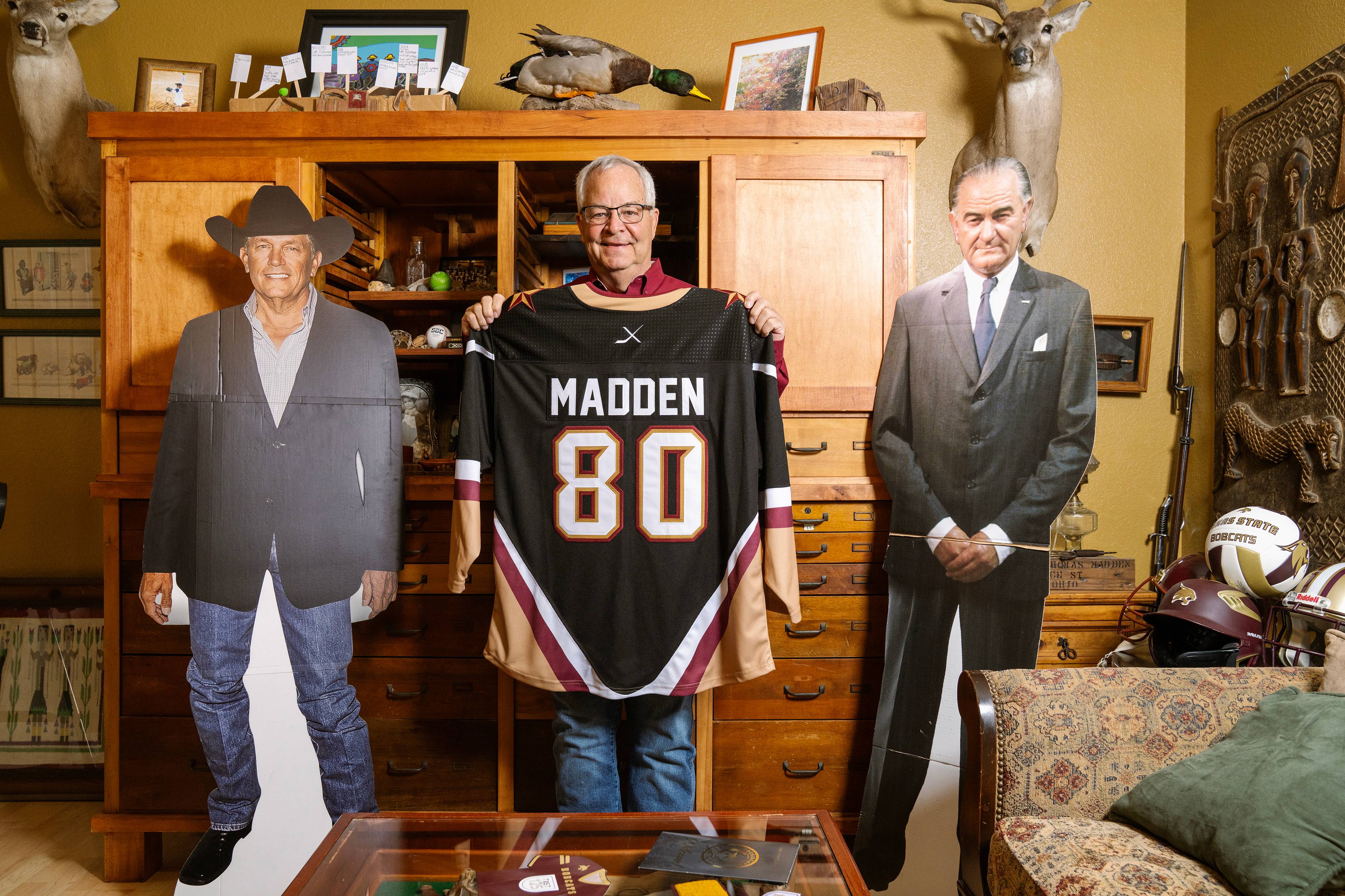 man holds up a hockey jersey with MADDEN on the back. he is standing between cardboard cutouts of LBJ and George Strait