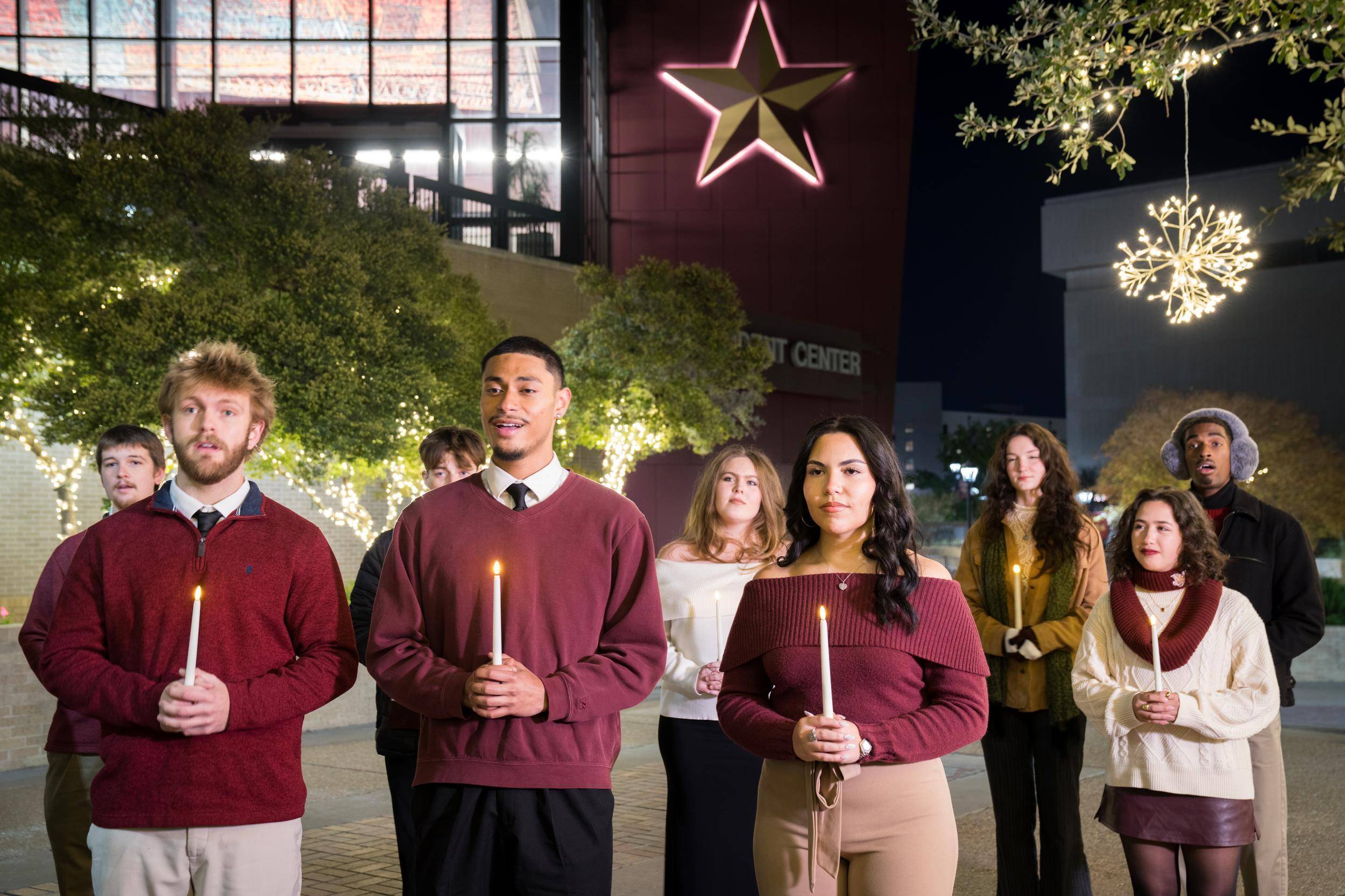 group of young adults in maroon and white winter attire holding up white taper candles while caroling