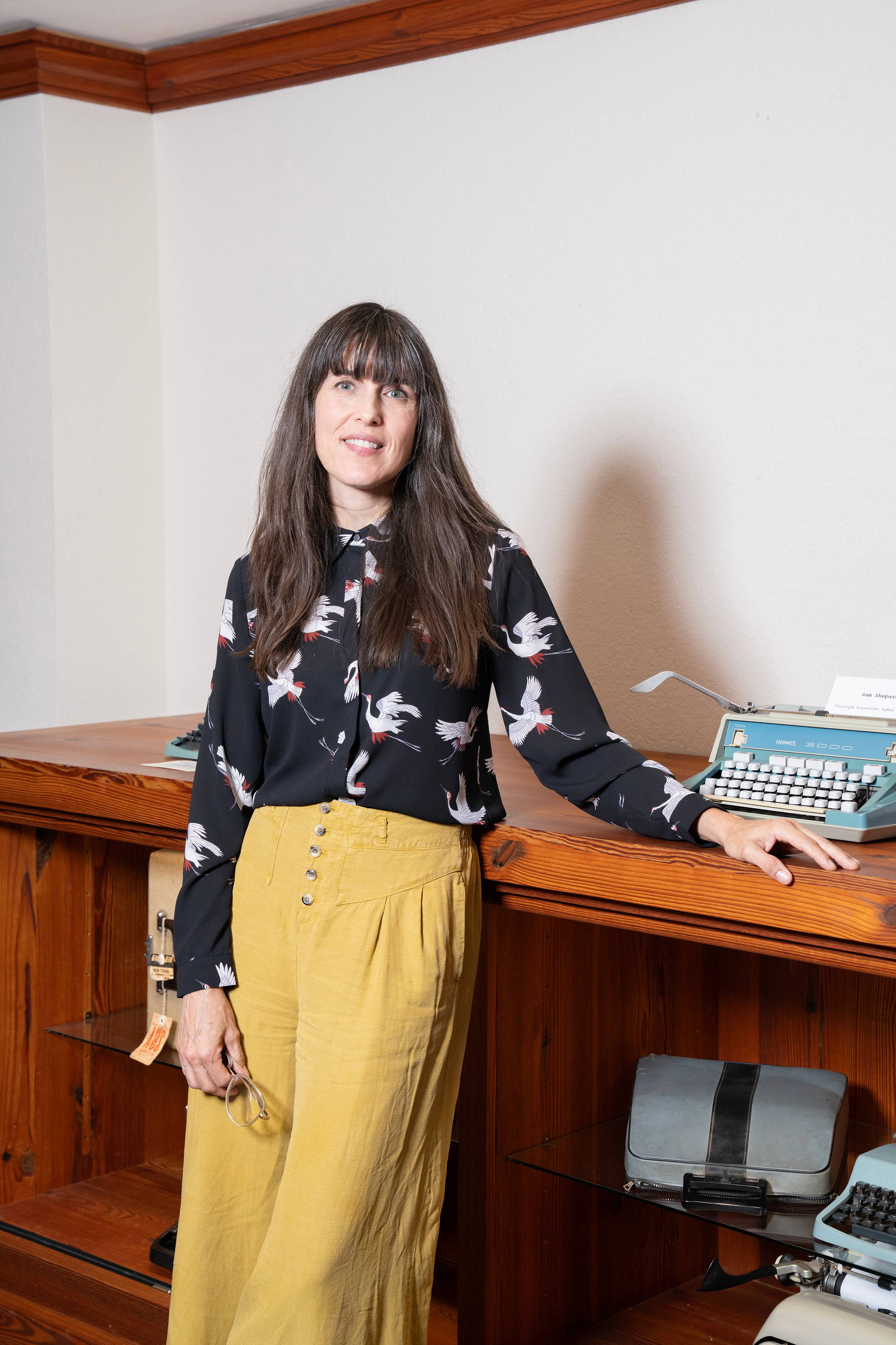 Carrie Fountain leans against a book case holding vintage typewriters