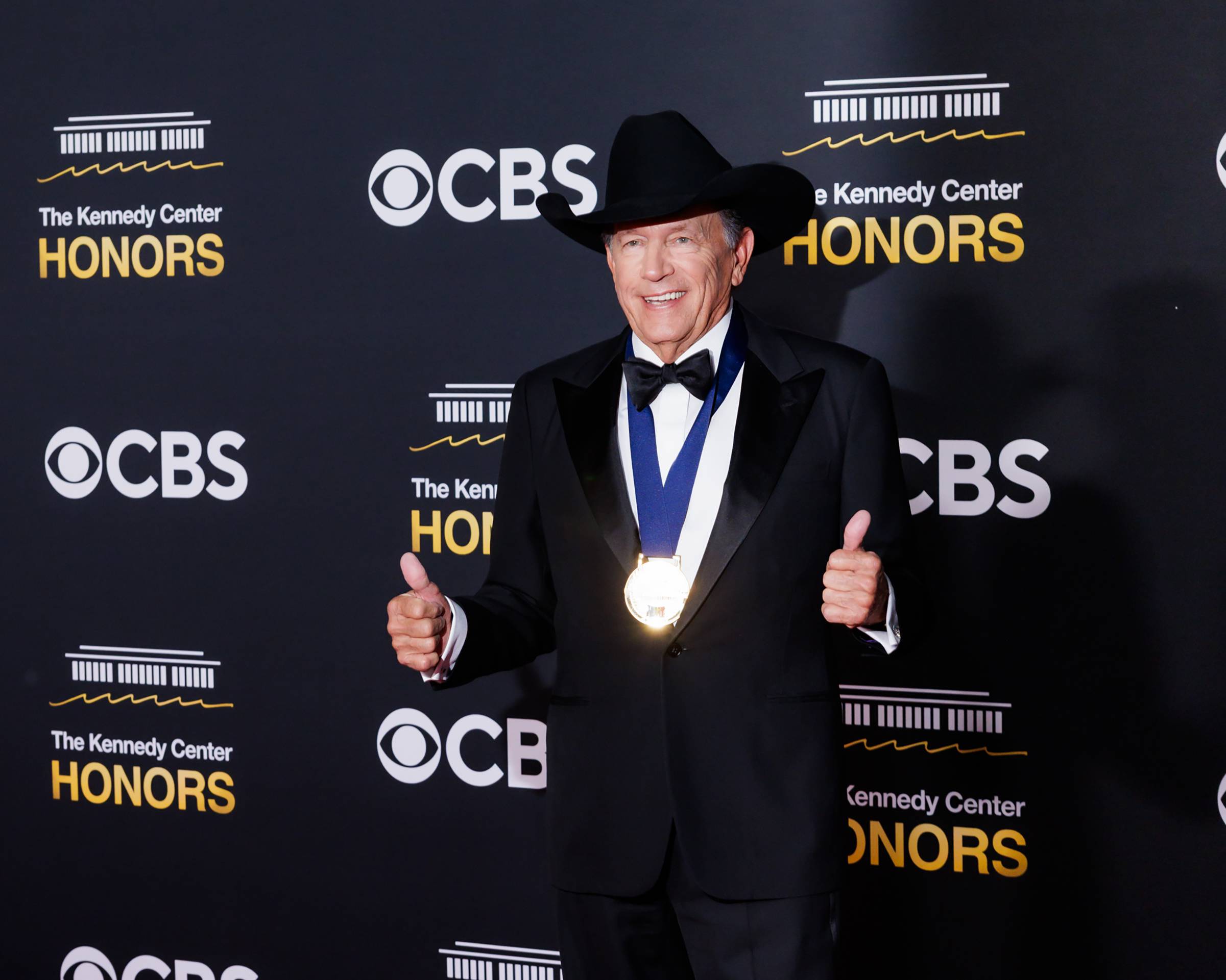 george strait stands in front of step and repeat wearing a tux and gold medal around his neck. both hands are giving a thumbs up