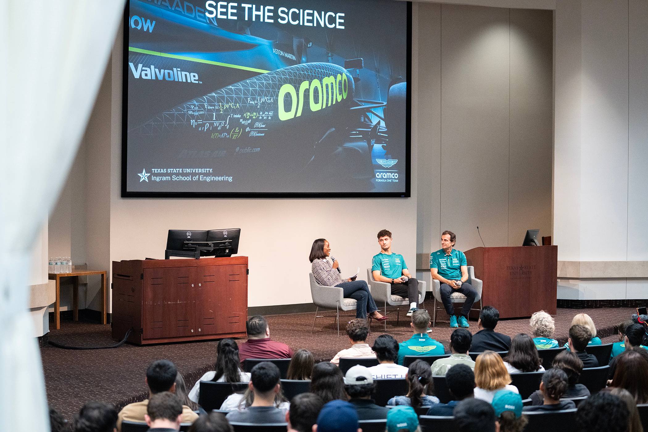 Wide angle of Formula One host Derin Adetosoye, drivers Pedro de la Rosa and Jak Crawford onstage in front of a crowd of students.