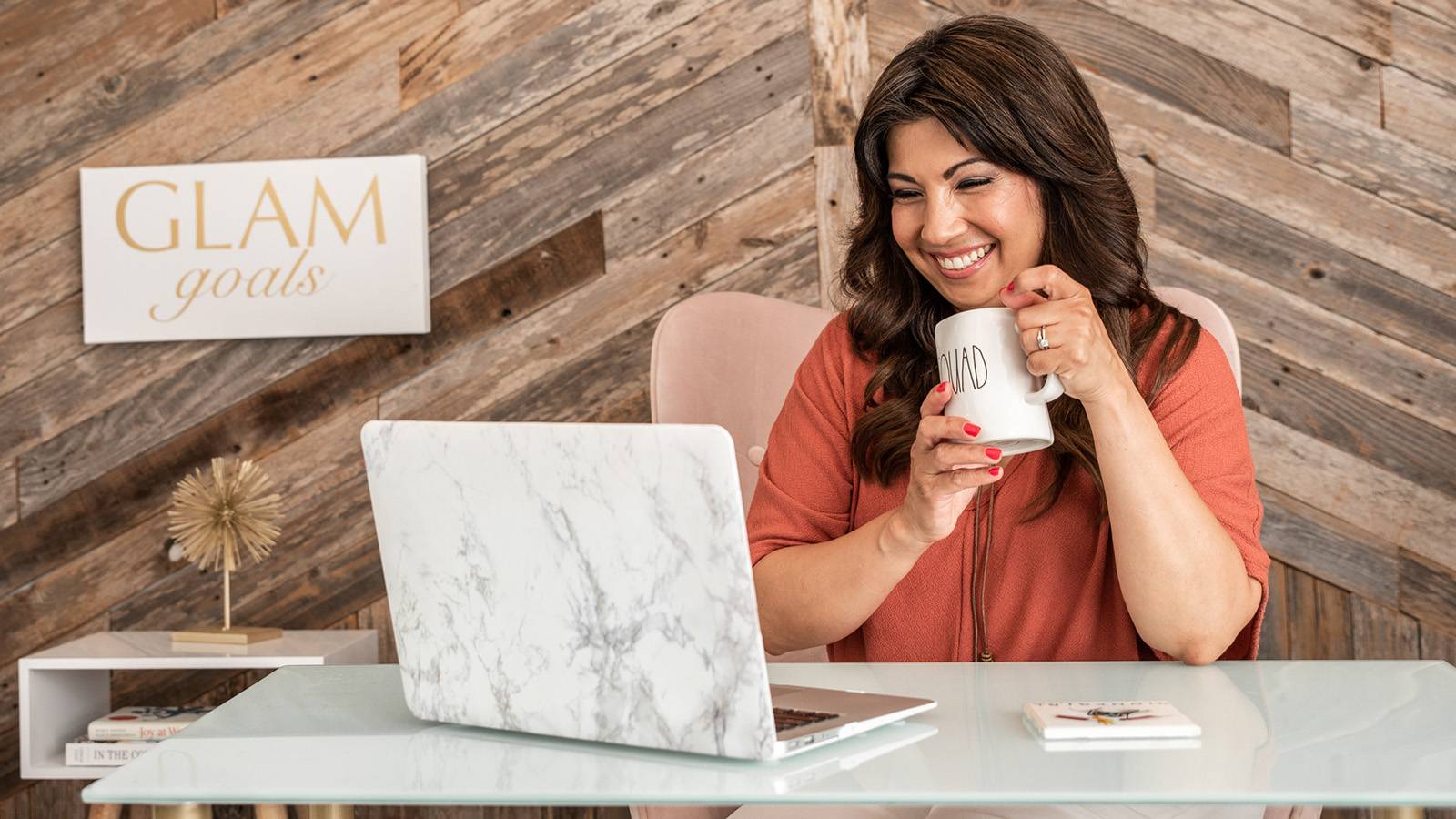 woman sitting at desk smiling while looking at laptop and holding a mug
