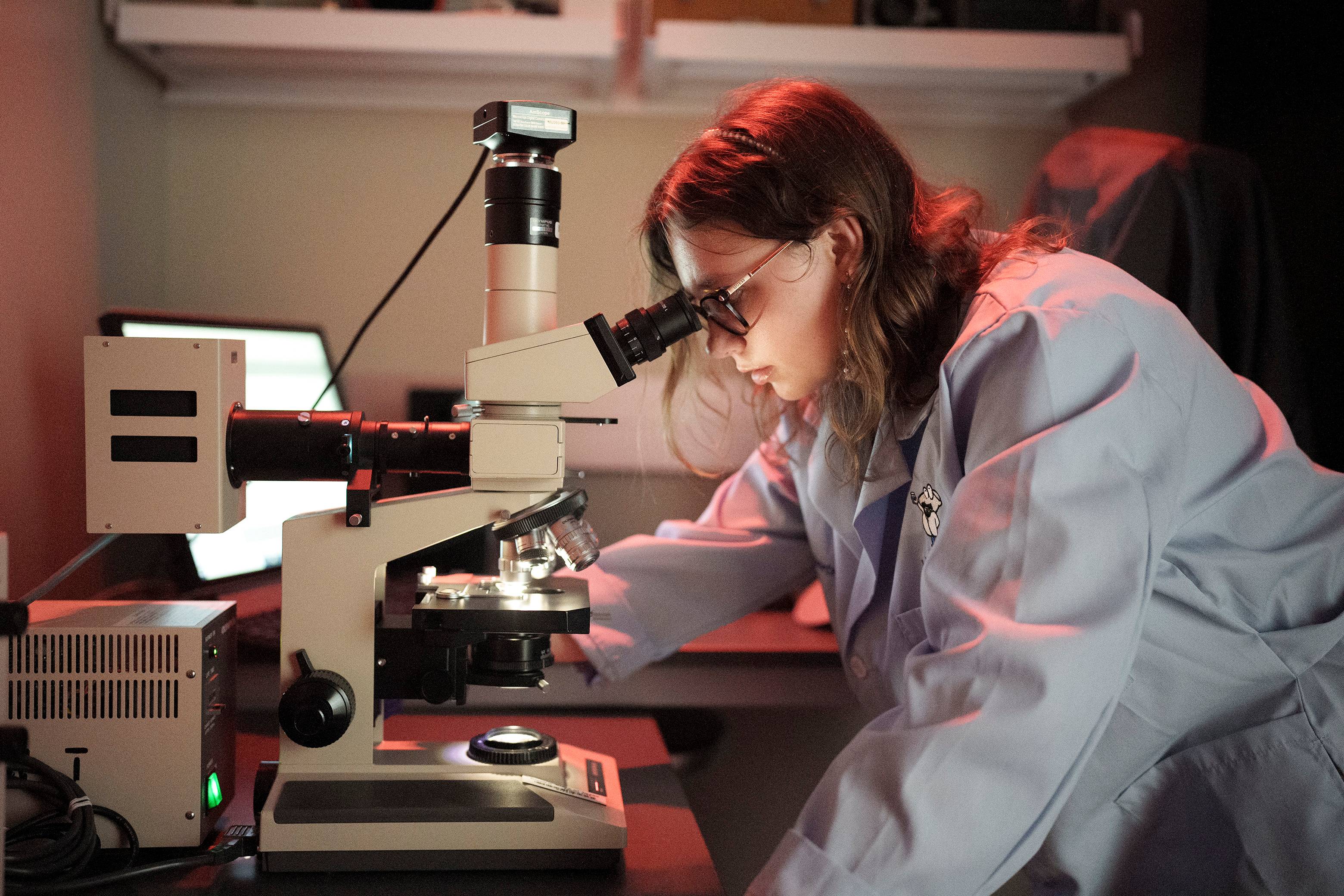 female student in white lab coat looks into microscope