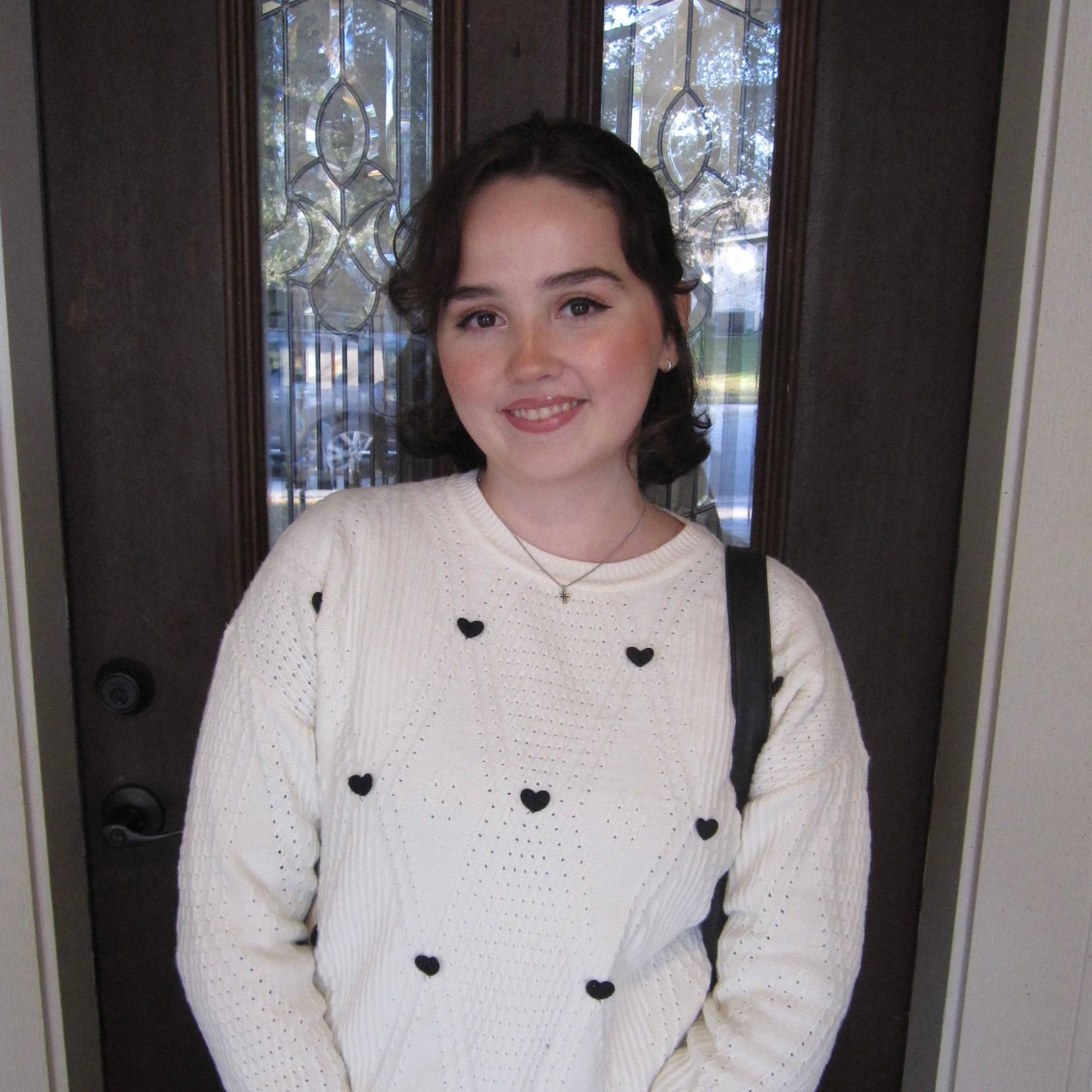 woman in white sweater with black hearts, bag strap over shoulder, door behind her