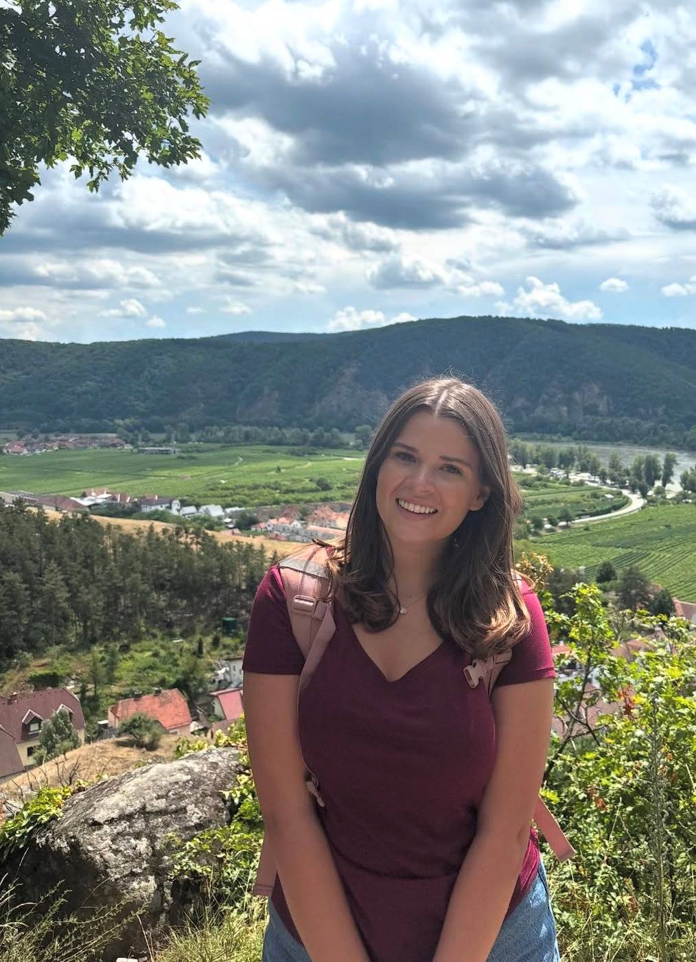 woman in red shirt, backpack, with river, mountains, and village behind her